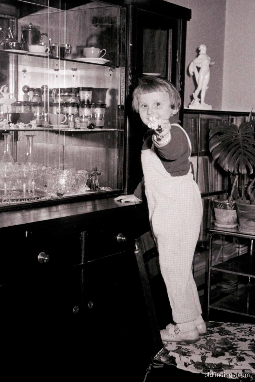 Young boy in corduroy overalls and a dark turtleneck playfully gestures with a finger, mimicking a shooting action. The photograph features a detailed interior scene with a large glass display cabinet and potted plants. Likely a 1960s-1970s family snapshot. Offers retro appeal.