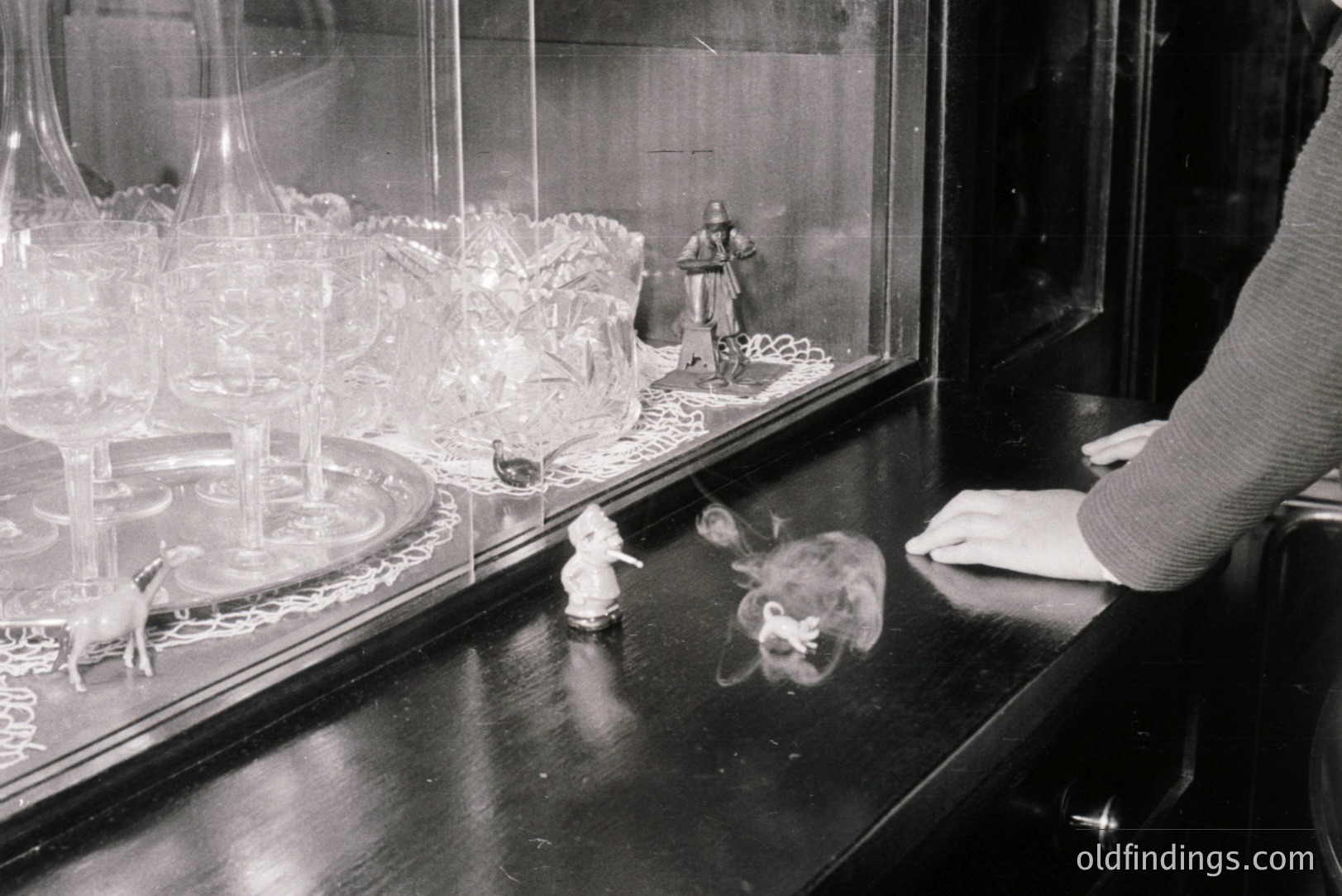Black and white photograph shows a glass display case with decorative figurines and cut-glassware. The scene includes a man in historical garb, miniature animals, and a fuzzy, indistinct object. A hand is partially visible on the case's edge. Likely a vintage shop or home display.
