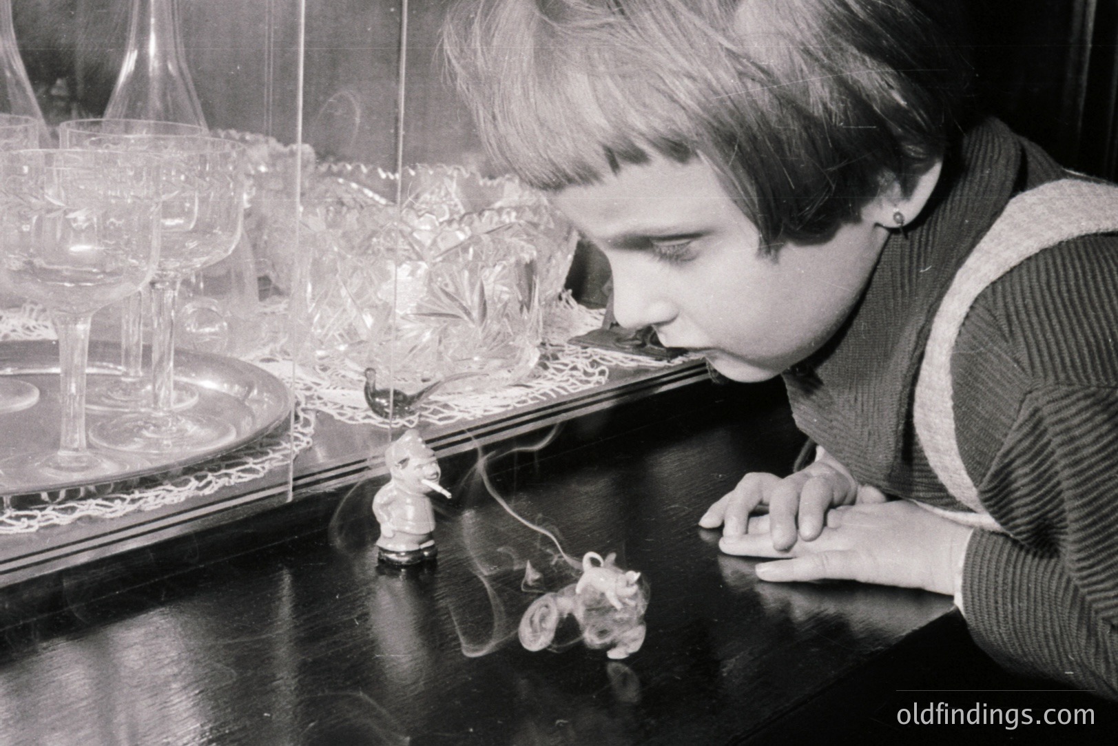 A young boy, profile view, intensely observes miniature figurines displayed behind glass. The scene suggests a museum or antique shop exhibit. Likely 1960s-1970s based on the boy's hairstyle and clothing style. The glass reflects multiple objects, creating an intriguing visual layer.