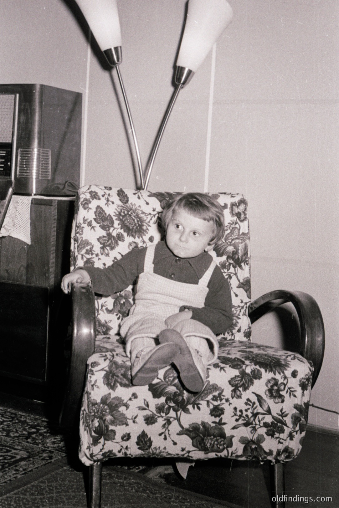 Young boy sits in a patterned armchair, wearing overalls and a dark sweater. A vintage floor lamp looms behind. Mid-century modern design evident in both chair and lamp style. Likely a candid portrait, c. 1960s-1970s American home. Stock potential for design & nostalgia.