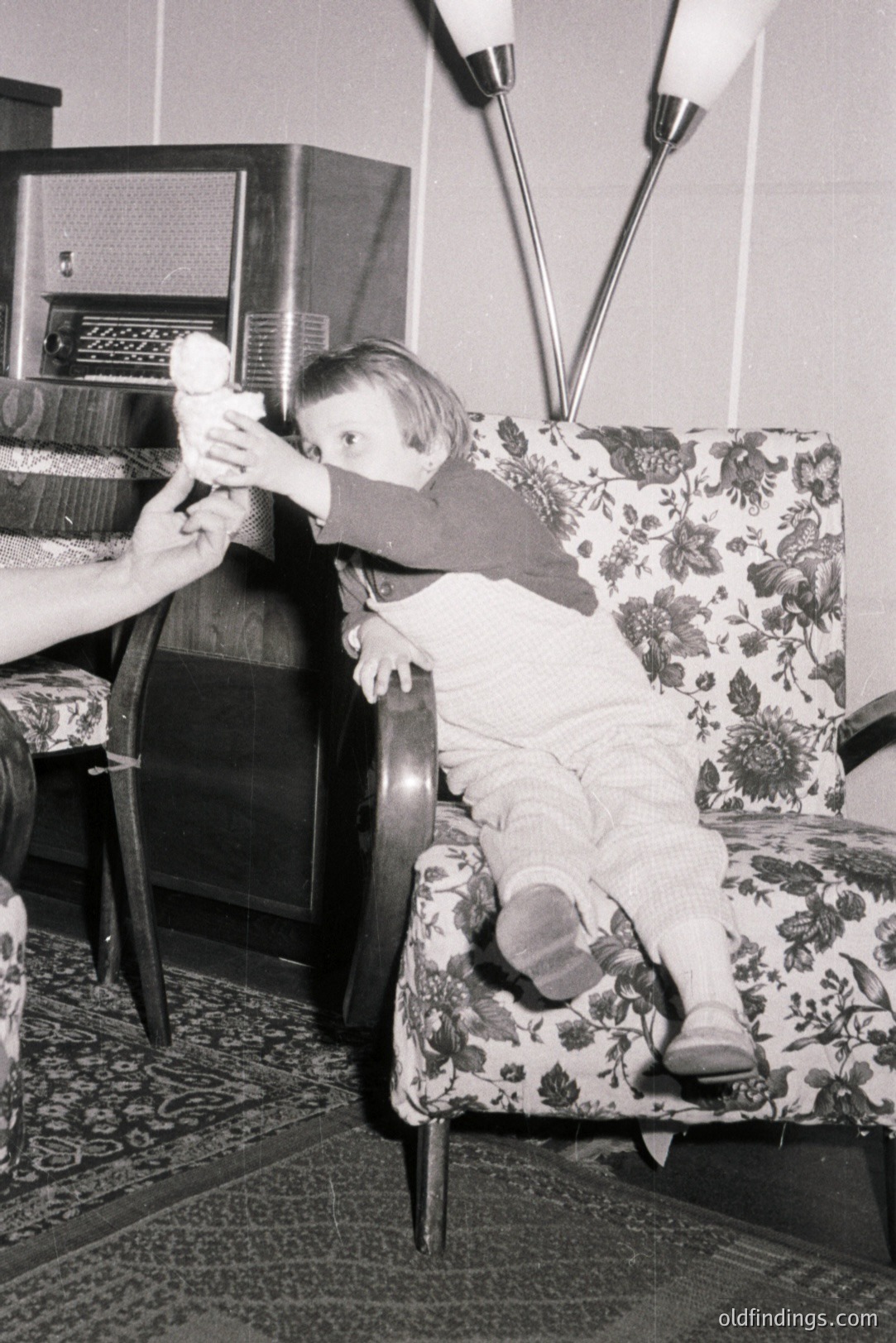 A young child, clad in a red sweater and patterned trousers, sits in a patterned armchair, reaching for an object held by an unseen adult. Vintage radio and a floor lamp establish a mid-century modern interior. Likely a family snapshot, c. 1950s-1960s. Captures domestic life and design trends.
