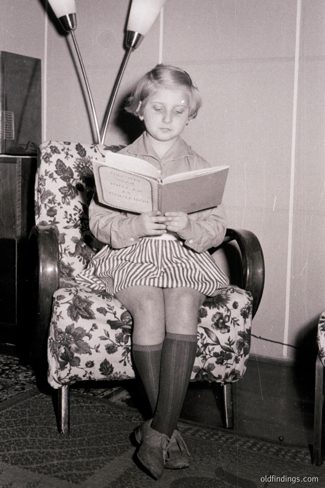 Young girl engrossed in a book while seated in a floral-patterned armchair. The decor suggests a mid-century interior with a distinctive floor lamp. Striped dress, knee socks & dark shoes complete the look. Likely 1950s. Stock photo potential for nostalgia themes.