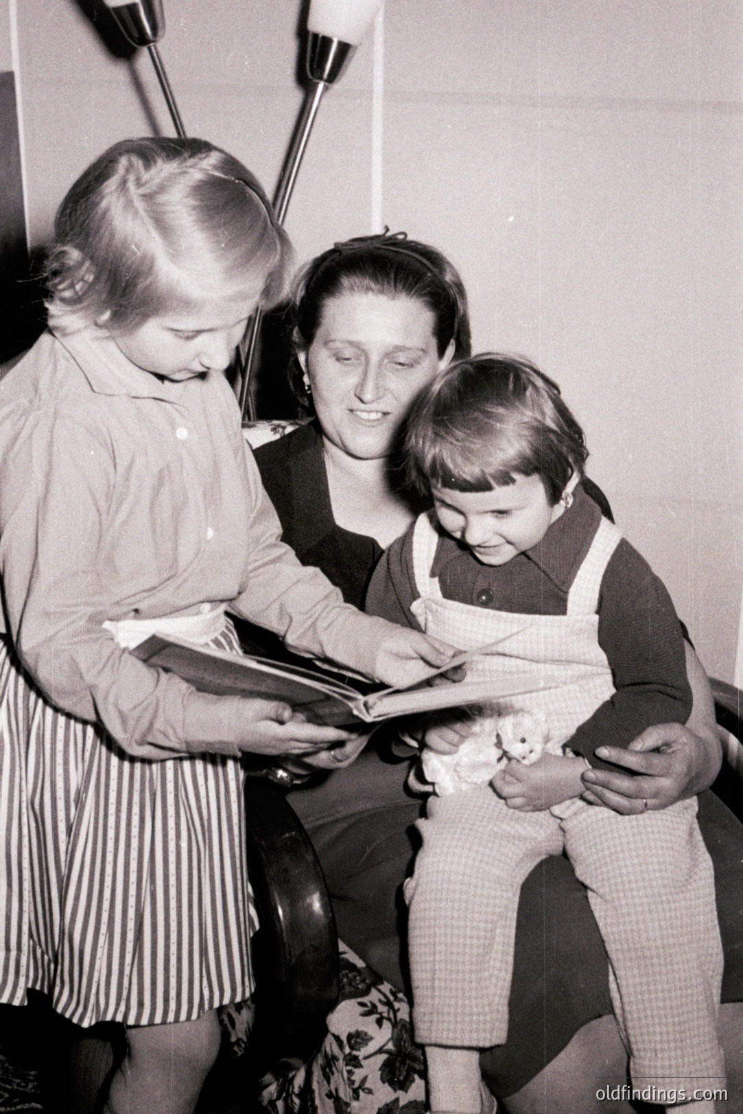 A mother sits in a patterned armchair, reading a book to two young children. Both children appear engaged, one standing and peering over her shoulder. A lamp is visible in the background. Likely a candid family moment, possibly 1960s or 70s. Domestic scene.