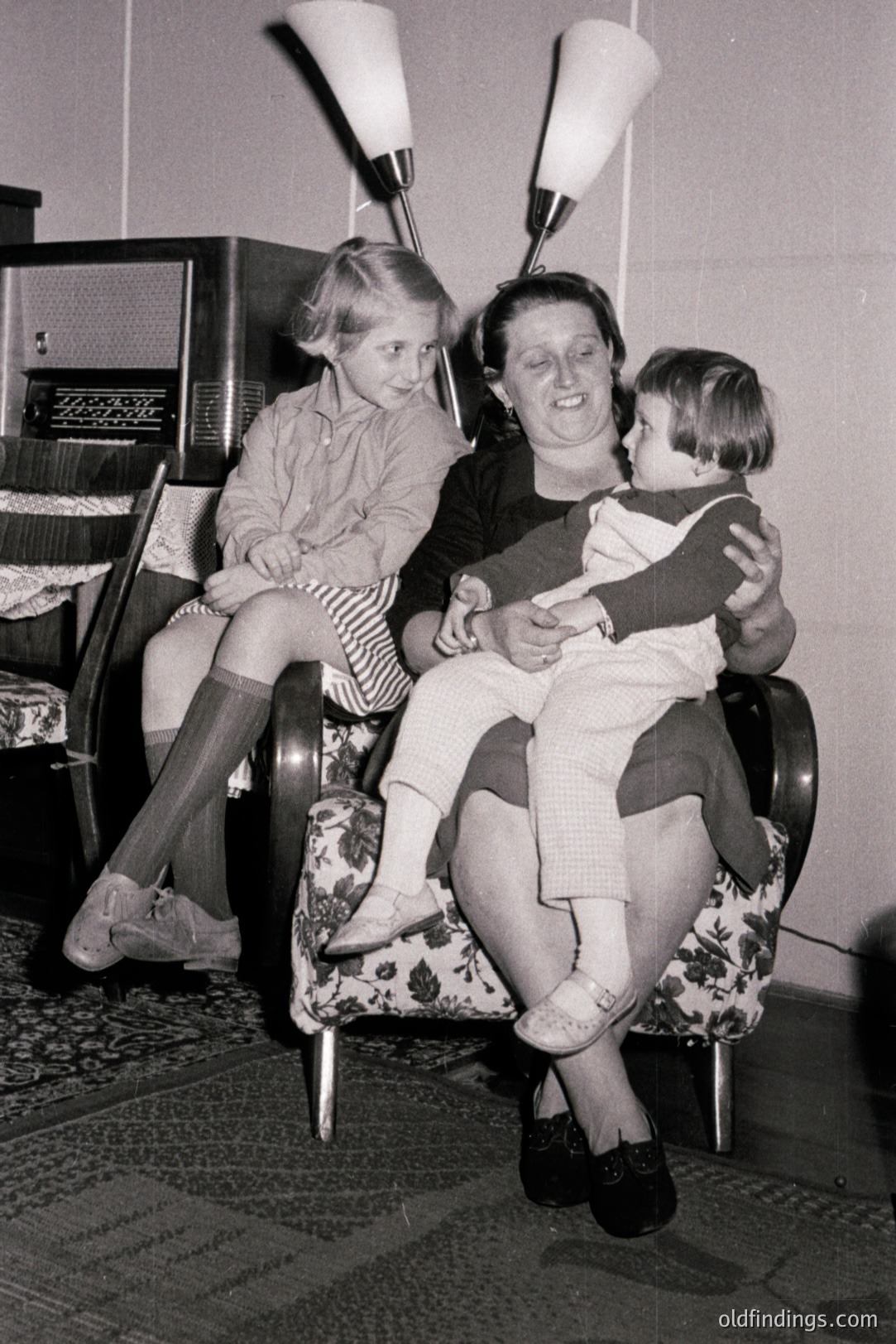 A mother cradles a baby in a patterned armchair, while a young girl sits nearby. Mid-century modern decor, including a radio and floor lamp, fills the room. Likely taken in the 1950s or 60s, this captures a domestic scene reflecting post-war family life. A nostalgic glimpse of everyday moments.