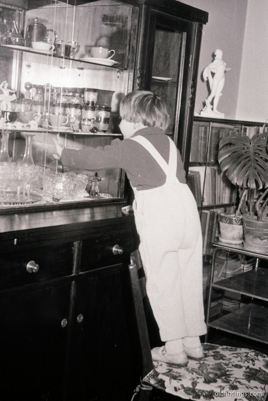A young child in overalls stands on a patterned chair, reaching towards a tall, ornate glass display cabinet. The cabinet holds numerous glass objects and figurines. A marble bust and potted plants are visible in the background. Appears to be an interior scene, likely 1960s-1970s.