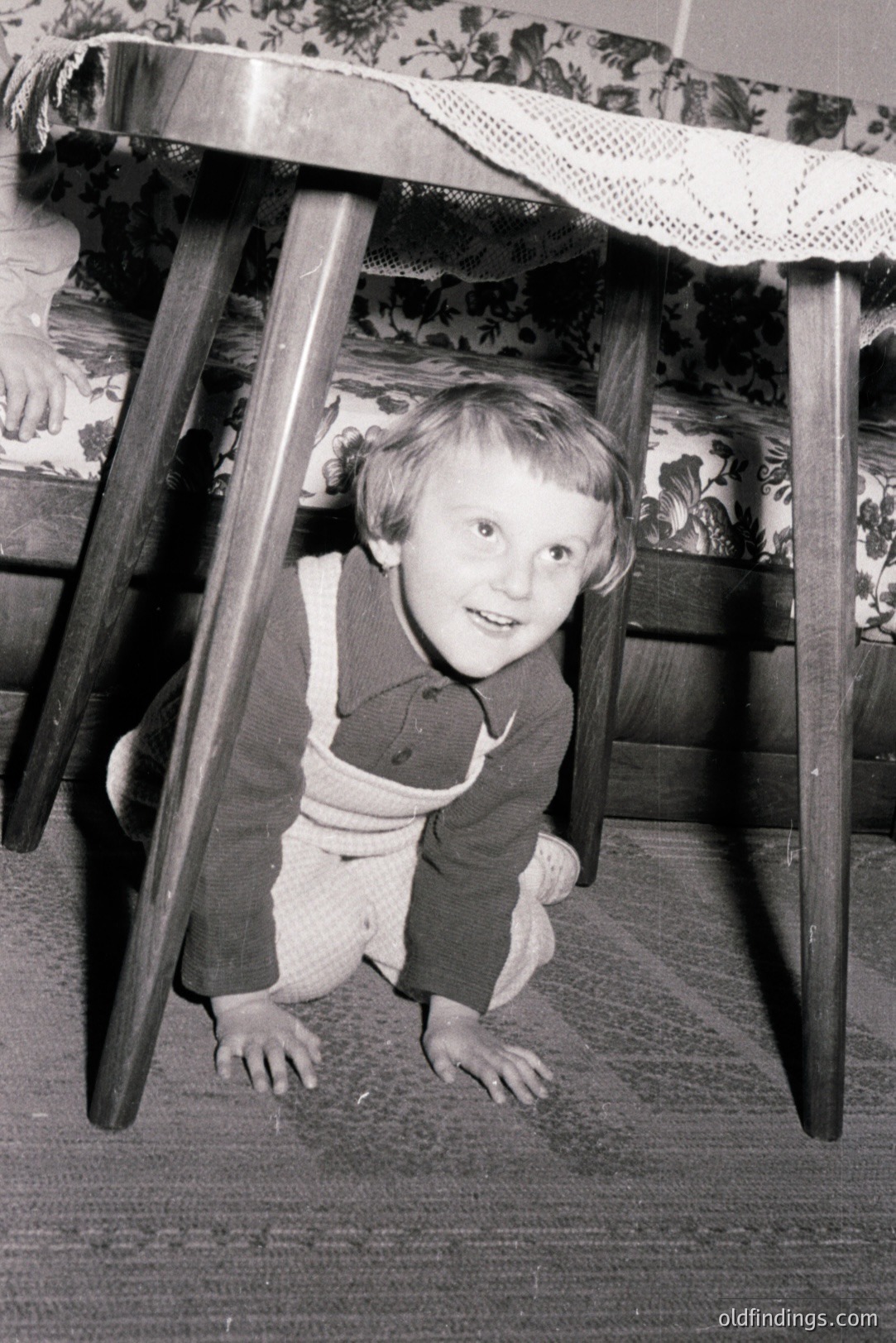 A young child crawls under a dark wood table with a patterned tablecloth, appearing to play hide-and-seek. The child wears a dark sweater and bib overalls. Floral patterned upholstery is visible behind the table. Likely a family snapshot from the 1960s or 1970s.