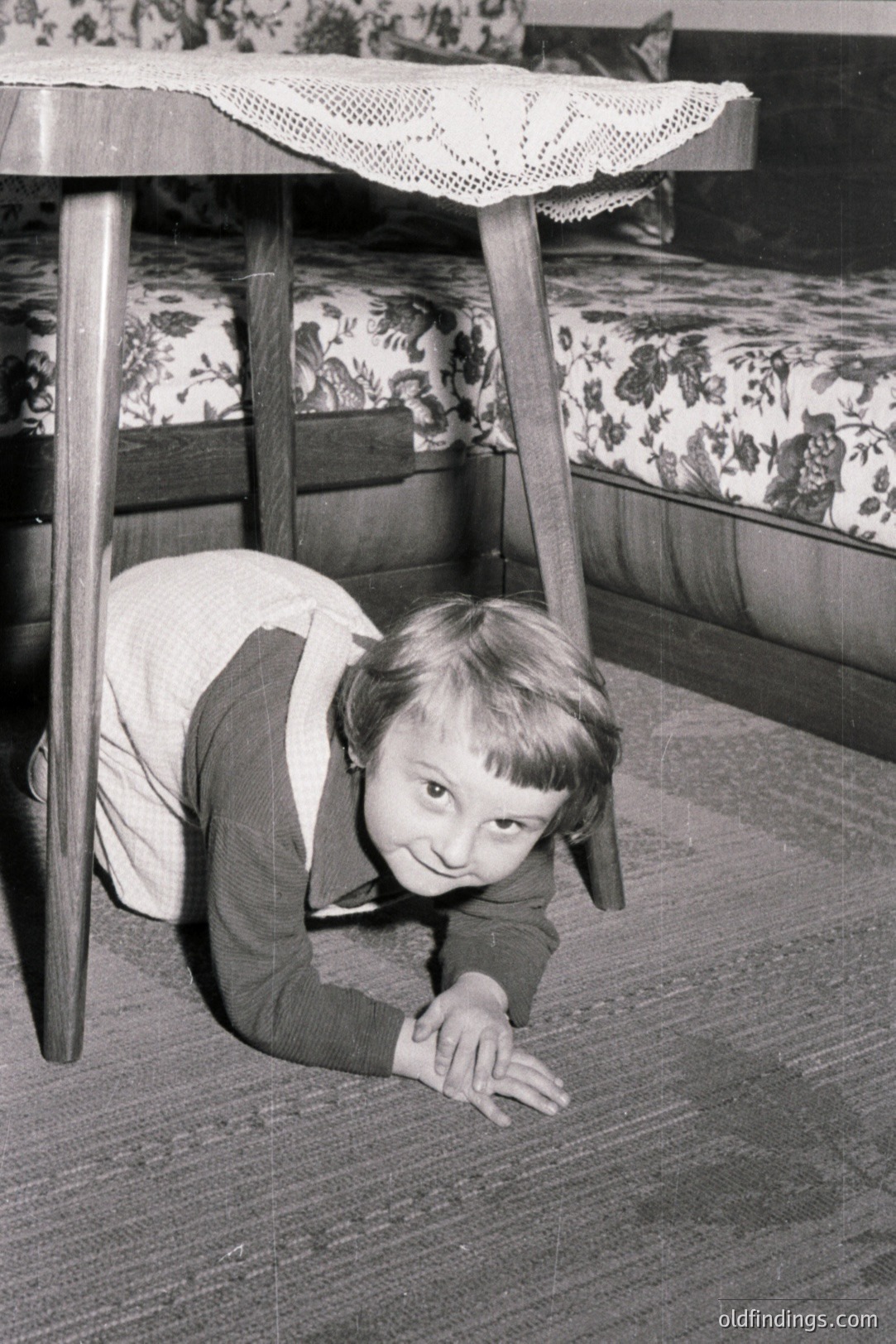 A child crawls playfully under a table with a lace tablecloth, showcasing a vintage domestic scene. The room features floral upholstered furniture. Likely taken in the 1960s or 70s, this image captures childhood games within a typical mid-century home.