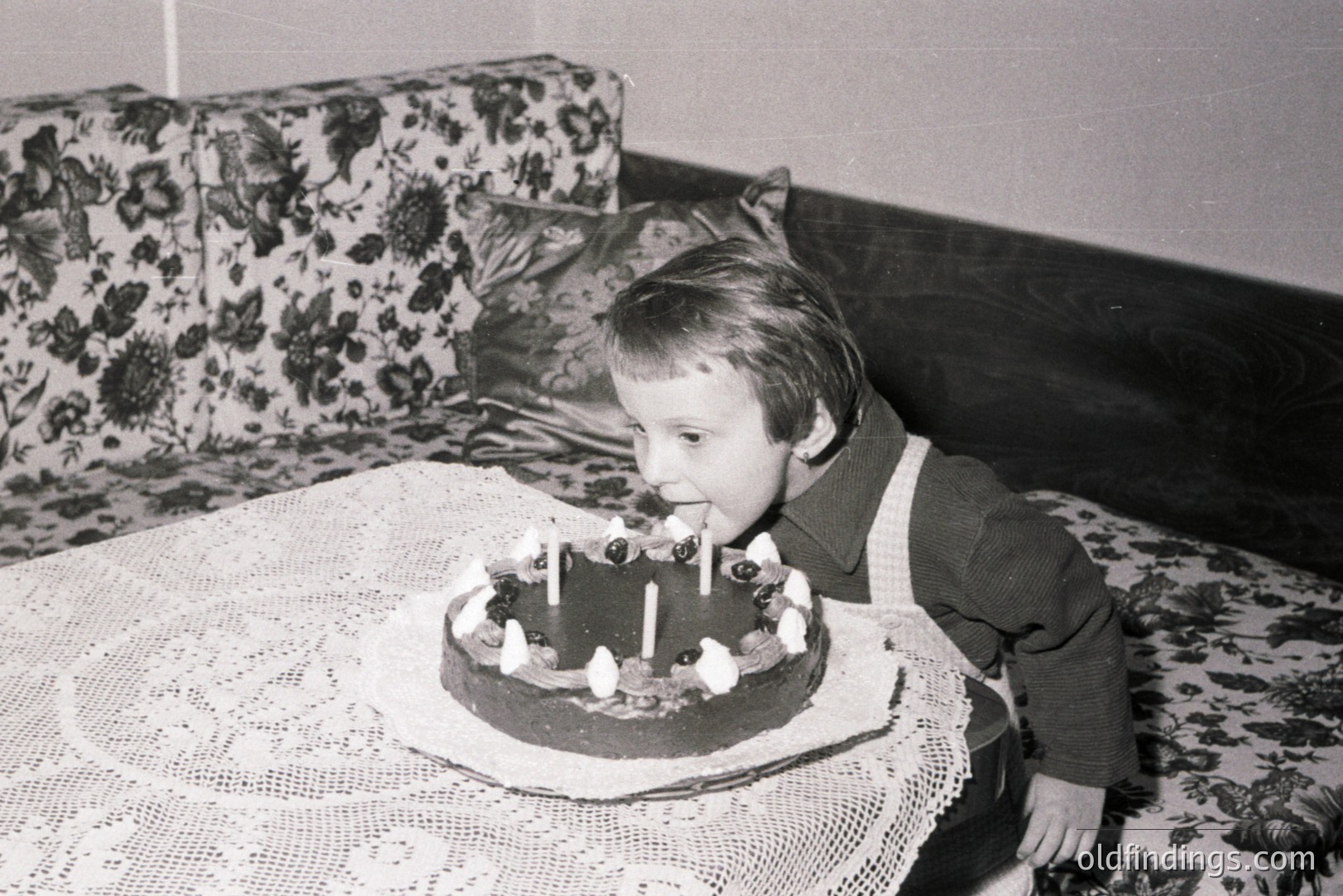 A young child, likely a boy, blows out candles on a chocolate cake adorned with cherries, seated on a floral-patterned sofa. A lace tablecloth covers the table. Likely taken in the 1960s or 1970s, capturing a family celebration moment. A typical domestic scene.