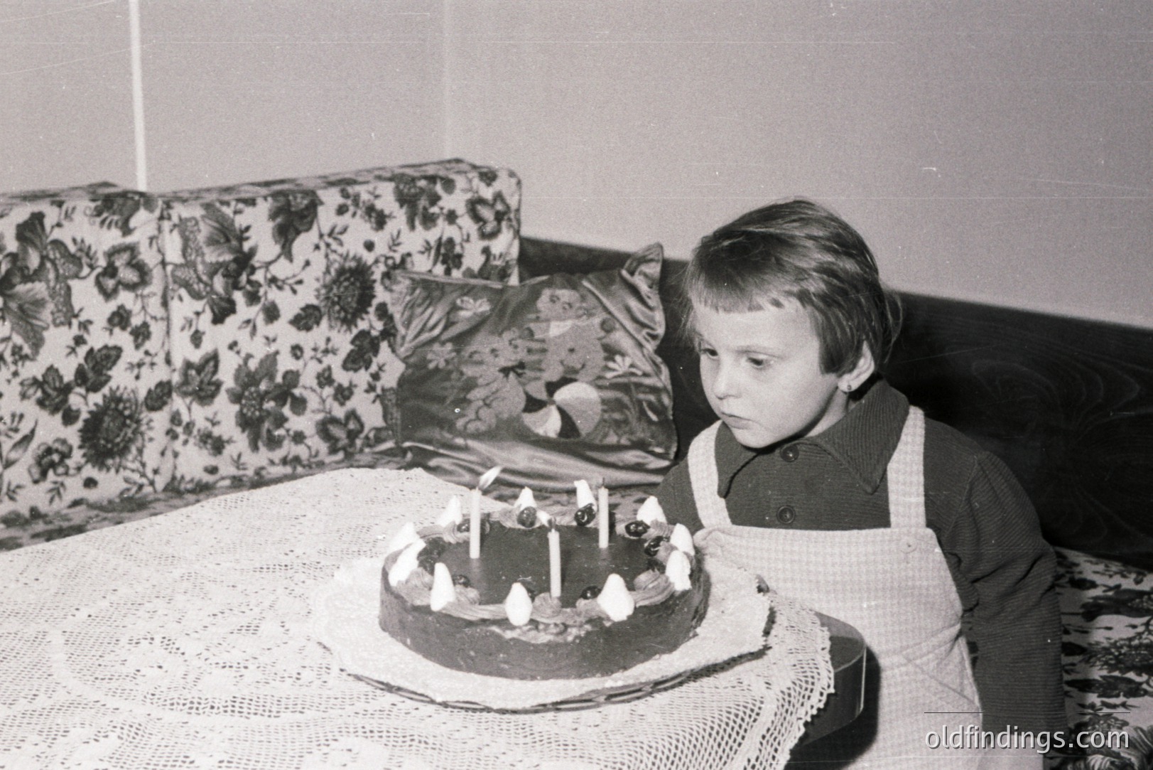 A young child, possibly a boy, stands observing a birthday cake with lit candles. The scene suggests a domestic setting, likely a home interior, with a floral-patterned sofa in the background. Appears to be a black and white photograph, indicative of the 1960s or 1970s. The child wears a collared shirt and apron.