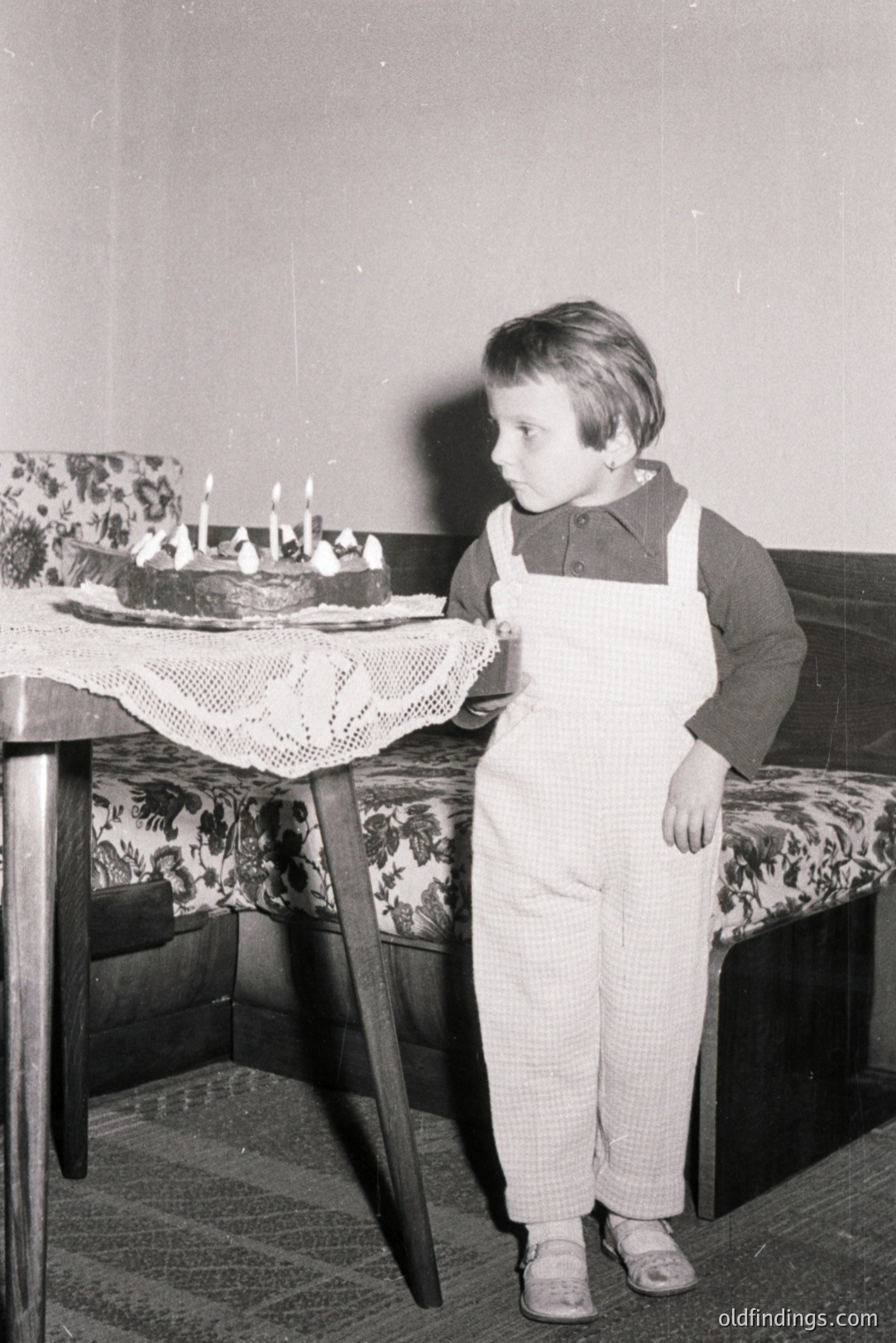 A young boy stands beside a table laden with a birthday cake, adorned with lit candles. He wears checked overalls over a dark shirt. The room features a patterned bench and table covered in lace. Likely a family gathering, c. 1960s. A glimpse into childhood celebrations.