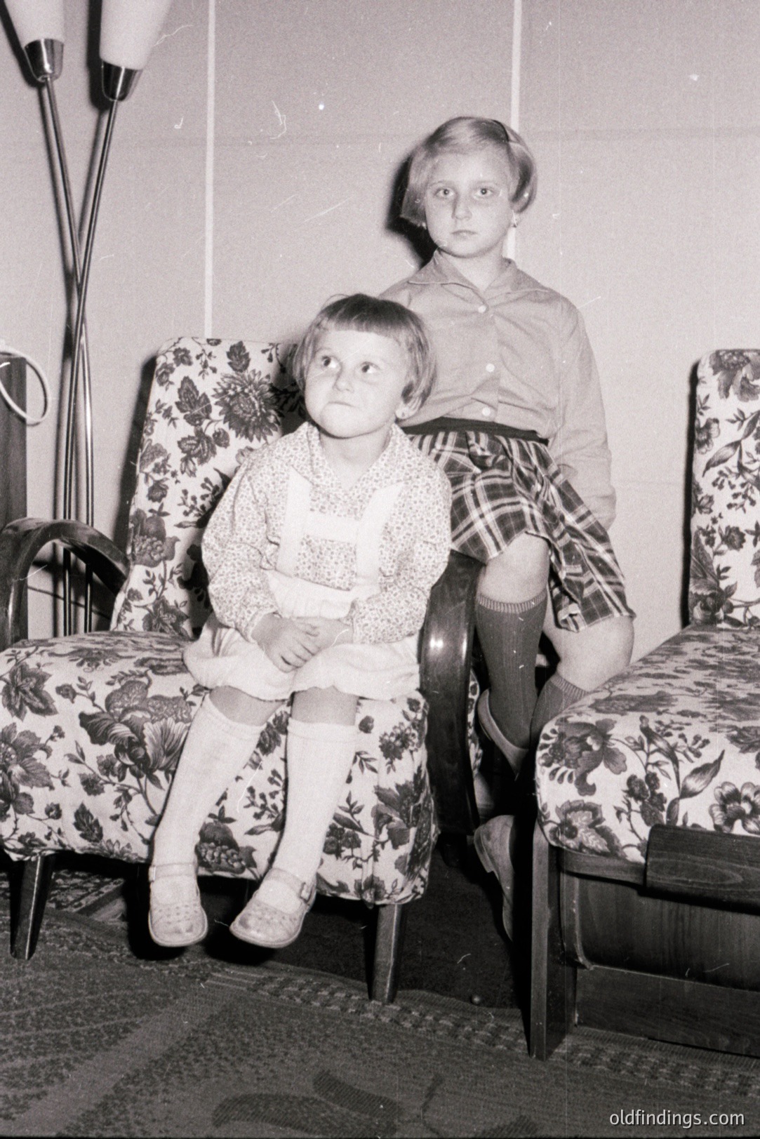 Two young girls seated in patterned armchairs, captured in a candid black and white snapshot. One wears a dress and knee socks, the other a collared shirt and plaid shorts. Likely a 1960s family portrait showcasing mid-century home décor. Simple, domestic scene. Possible stock photography reference.