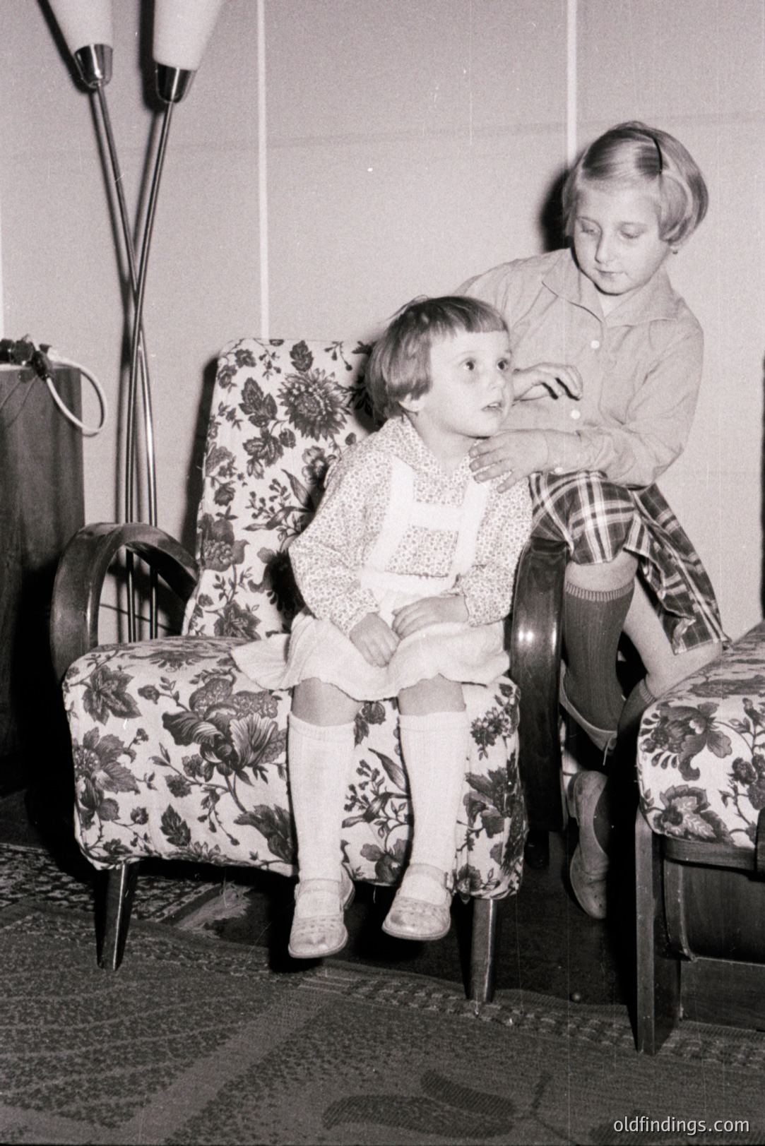 A young girl in a floral-patterned dress and tights sits in an upholstered armchair, with another child standing behind her. A floor lamp with a unique design stands nearby. Likely a domestic interior, reflecting mid-century design aesthetics. Possibly 1960s-1970s.