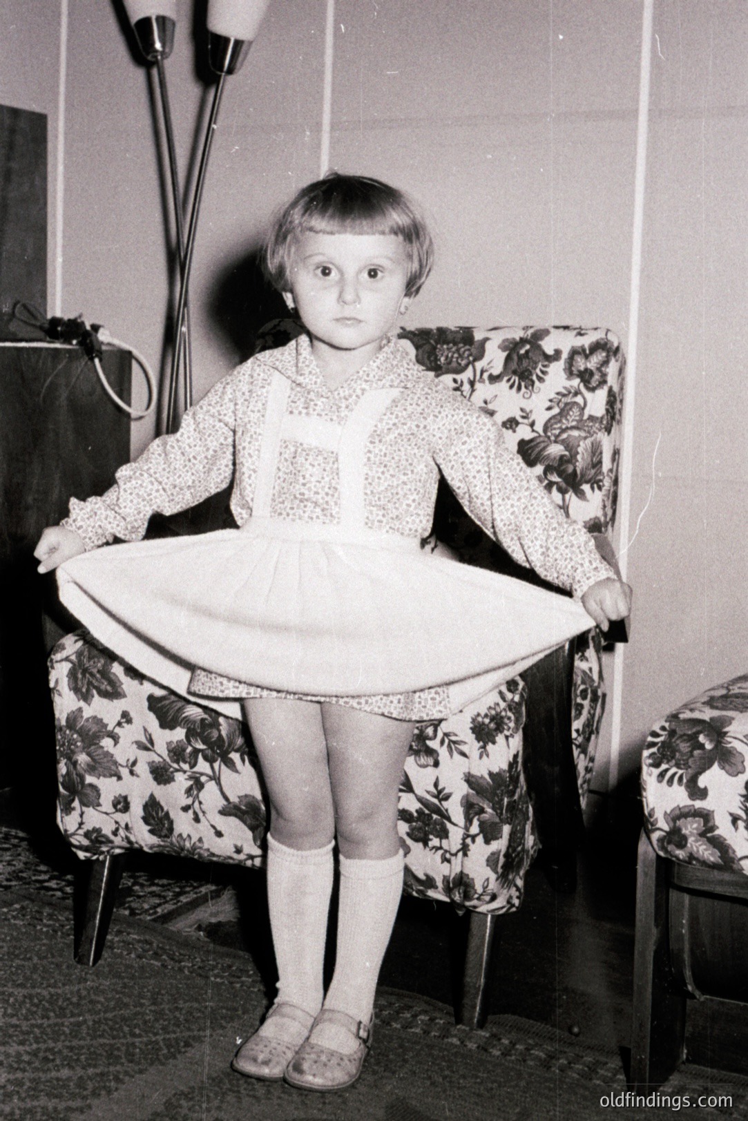 Young girl playfully holds up a dress, showcasing a frilled hem. She wears a patterned top, suspendered skirt, knee socks, and Mary Jane shoes. The room features floral patterned furniture and a tall floor lamp. Likely a domestic snapshot, mid-20th century.