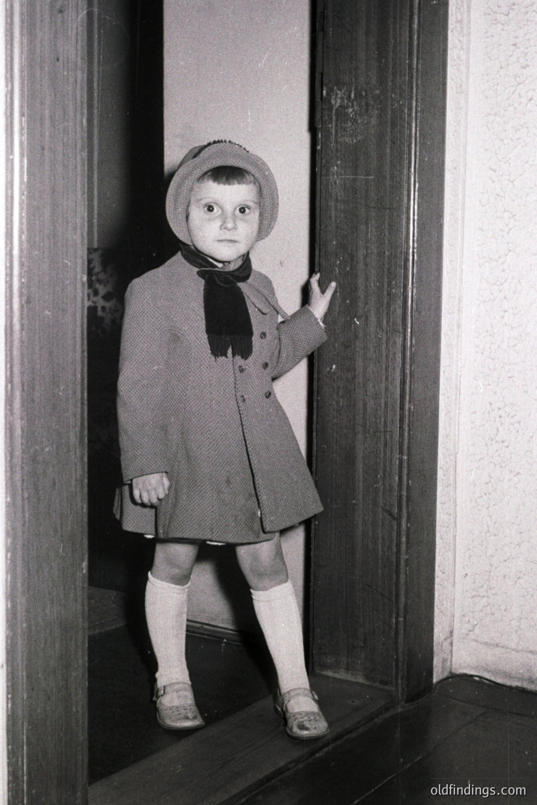 A young child, likely a girl, stands in a doorway wearing a wool coat, cloche hat, and saddle shoes with stockings. The scene appears candid, possibly a family snapshot. Architectural details suggest a modest, older home. Likely taken in the 1930s-1950s. Potential for historical research/family archives.
