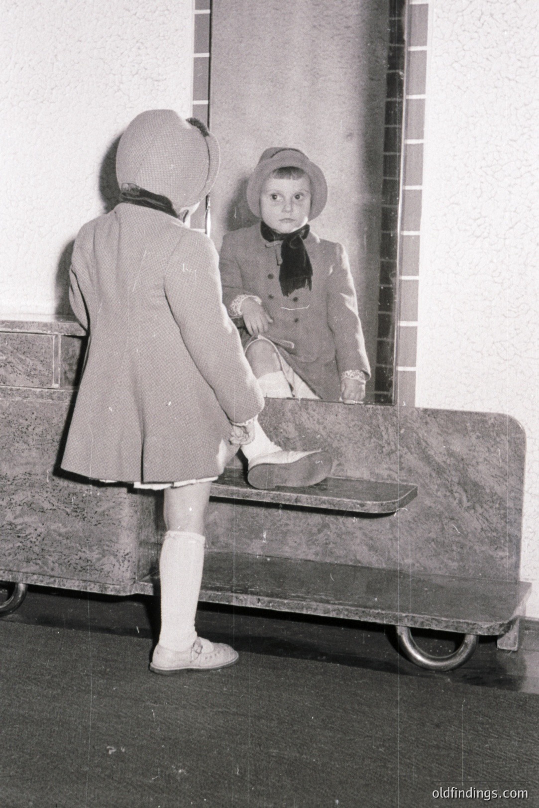 Young child in a double-breasted coat, hat, and knee socks, captured in a mirror reflection. Appears to be dressing or adjusting footwear. Likely a candid portrait, possibly a family snapshot, c. 1950s-1960s. Architectural details suggest a public space like a restroom or hall.