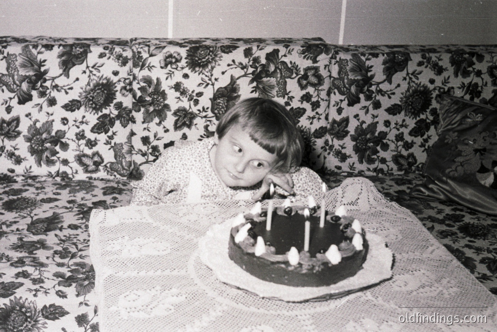 A young boy with a bowl cut smiles next to a chocolate cake with eight lit candles. He reclines against a floral-patterned sofa, covered by a vintage lace tablecloth. Likely a childhood birthday celebration, suggestive of the 1960s or 70s. Nostalgic family moment.