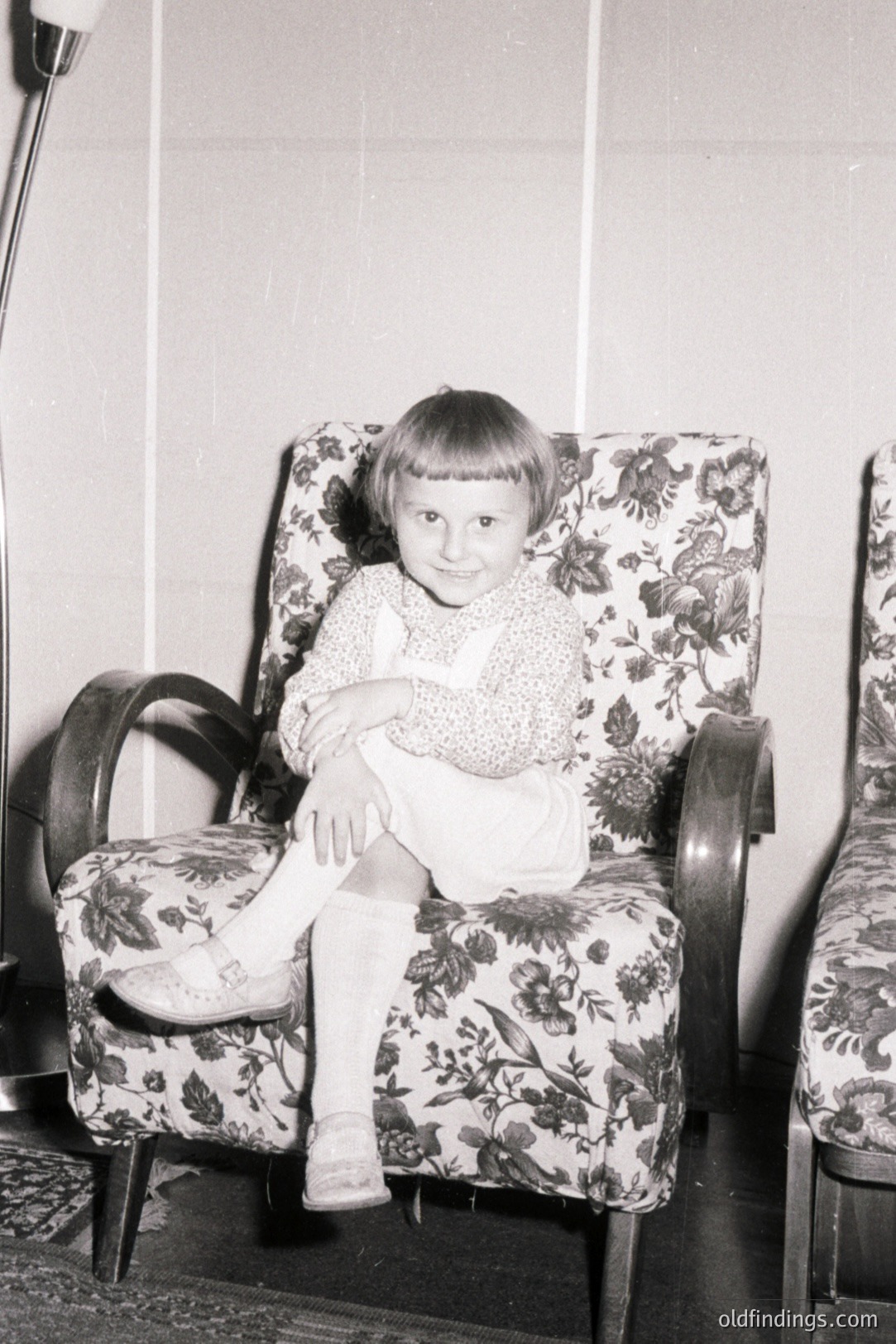 A young child sits formally in a patterned armchair, appearing to pose for a portrait. The room features paneled walls and a glimpse of a patterned rug. Likely taken in the 1950s or 1960s, demonstrating mid-century interior design. Offers visual references for period styling.