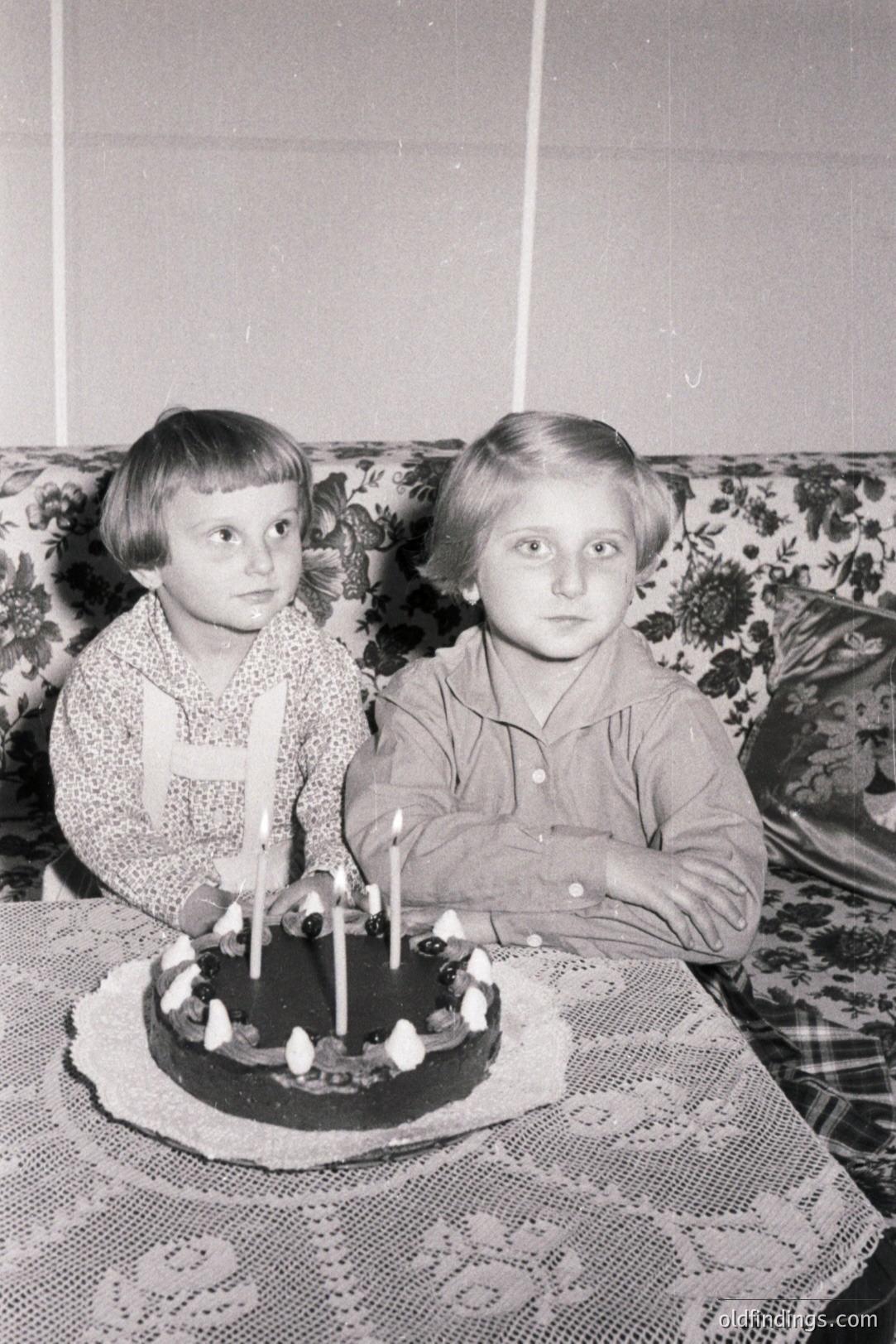 Two young children, a boy and a girl, sit side-by-side with a birthday cake featuring lit candles. The girl wears a collared shirt; the boy, a patterned button-down. Floral wallpaper and a patterned tablecloth frame the scene. Likely 1960s-1970s family snapshot. Nostalgic domestic imagery.