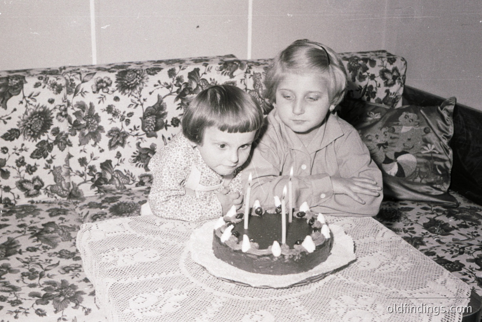 Two young girls celebrate a birthday with a chocolate cake adorned with seven candles. The scene appears domestic, set against a floral patterned sofa. Likely a family snapshot, indicative of mid-20th century home décor and childhood celebrations. Appears to be a snapshot.