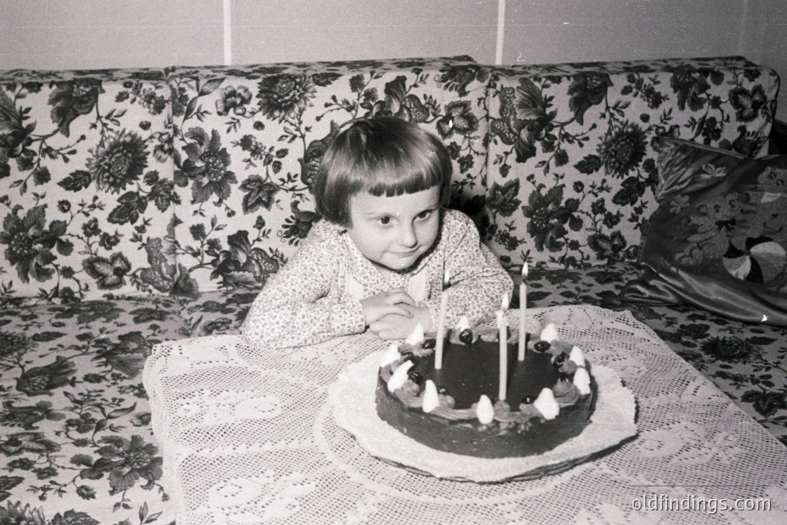 A young child, likely a girl, sits poised before a birthday cake with eight lit candles on a lace-covered table. The backdrop features a floral-patterned sofa. Likely a snapshot from the 1960s or 70s, capturing a domestic, celebratory moment. Image holds potential for nostalgia-themed design.