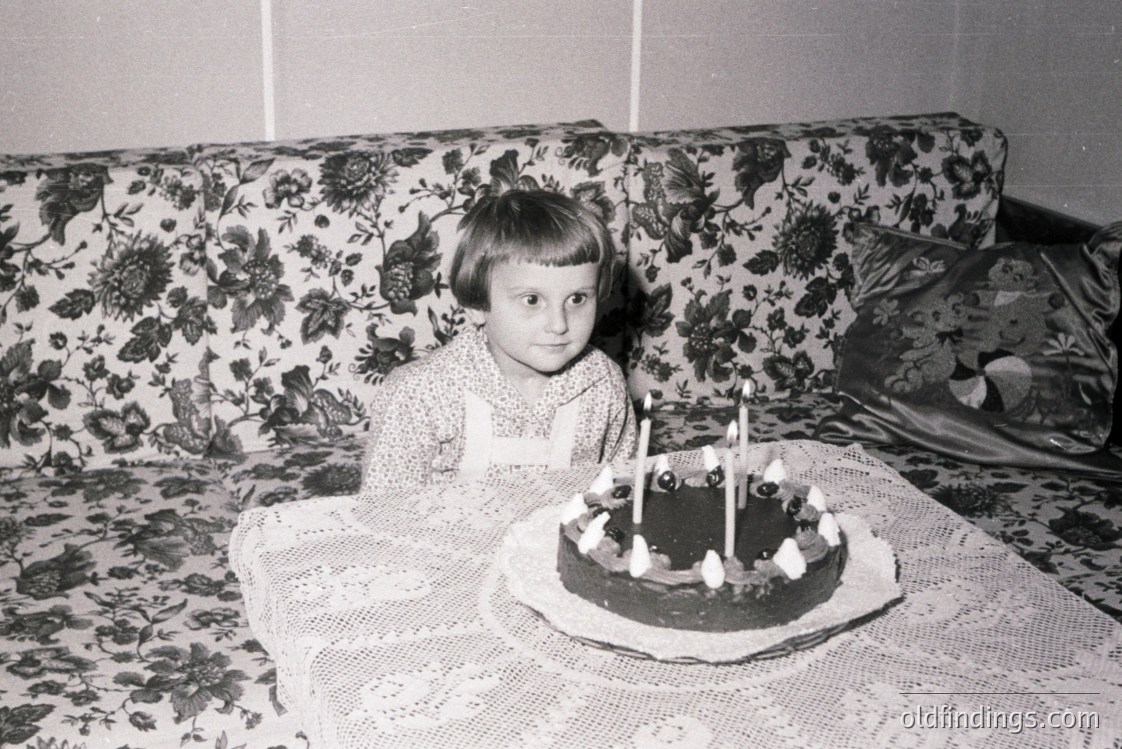 A young girl with short, dark hair sits behind a table adorned with a lace tablecloth, celebrating a birthday with a chocolate cake and lit candles. Background features a floral-patterned sofa. Likely mid-20th century, possibly 1960s, home interior. Nostalgic family moment.