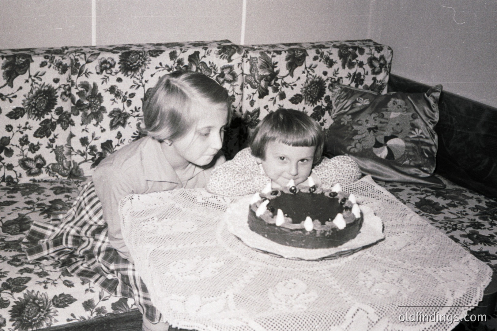 Two children celebrate a birthday with a chocolate cake adorned with seven candles. Interior scene features a patterned floral sofa and a lace-covered table. Likely mid-20th century, possibly 1950s-1970s. Captures a candid family moment, potential for nostalgia marketing.