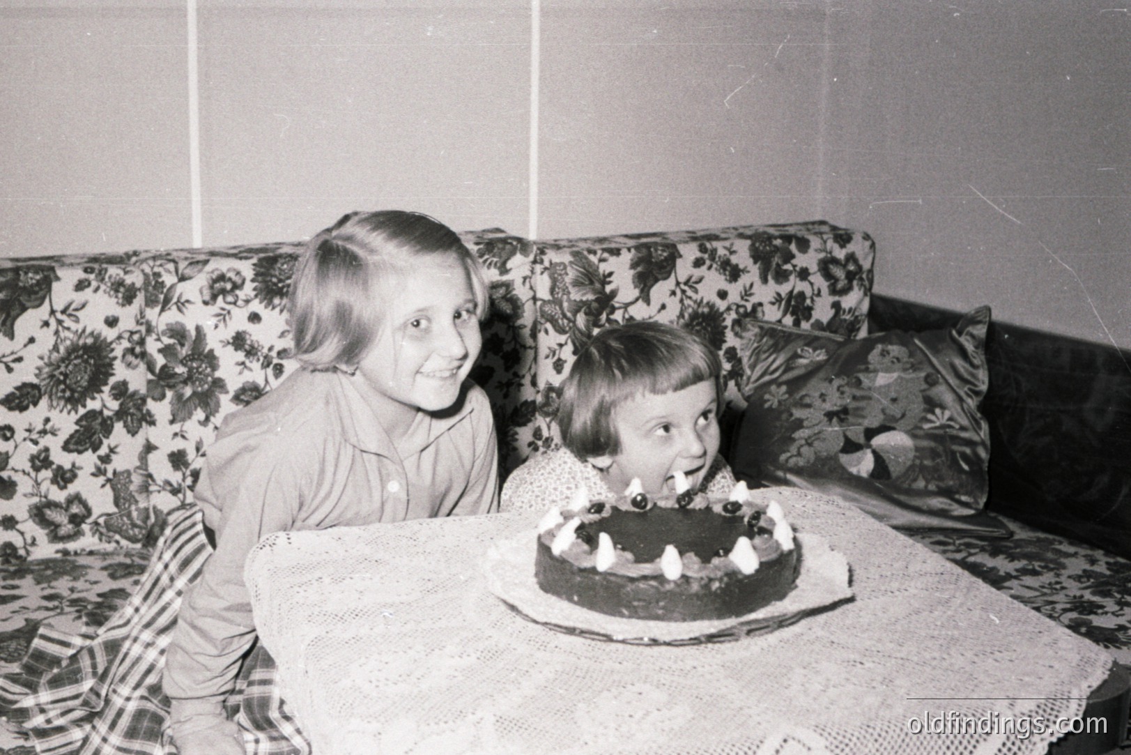 Two young girls share a moment of joy, excitedly observing a chocolate cake with lit candles. The setting appears to be a home interior, with a floral-patterned sofa as backdrop and a lace-covered table. Likely a family celebration, mid-century aesthetic.