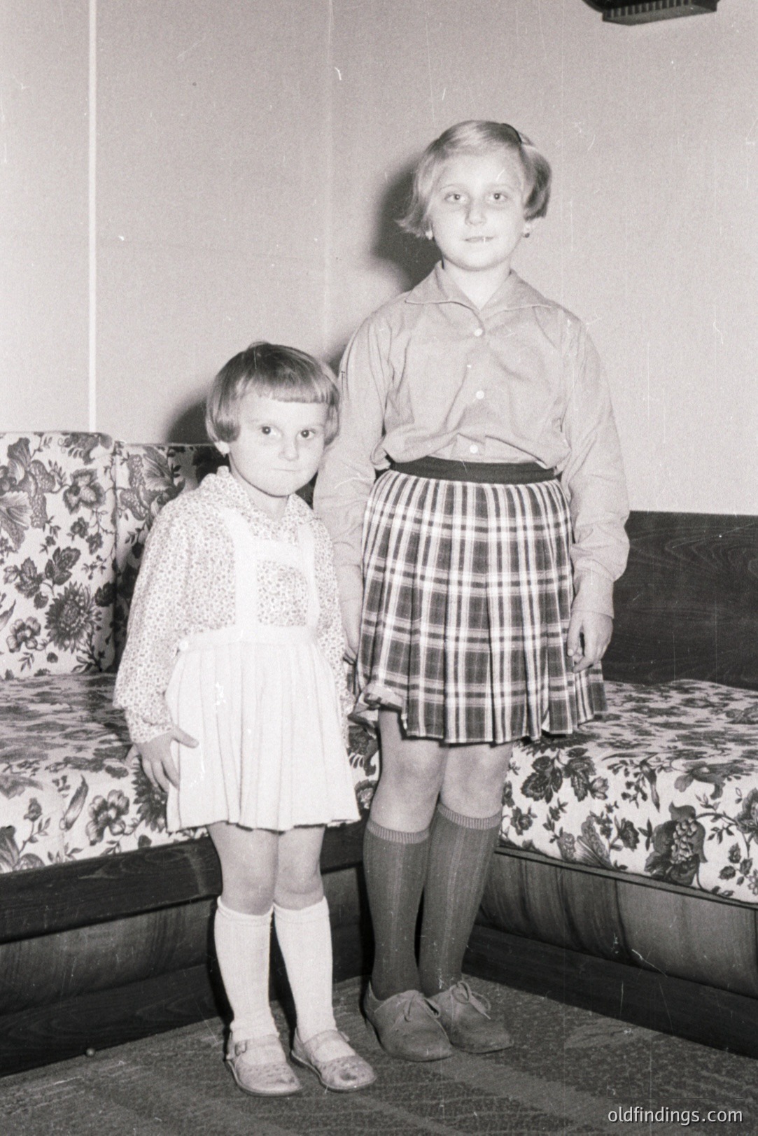 Two young girls stand near a floral-patterned couch. One wears a white, short-sleeved dress with lace trim; the other, a plaid skirt, collared shirt, and knee socks. Likely mid-century home interior, captures childhood fashion trends. Subject matter suitable for vintage design or family history research.