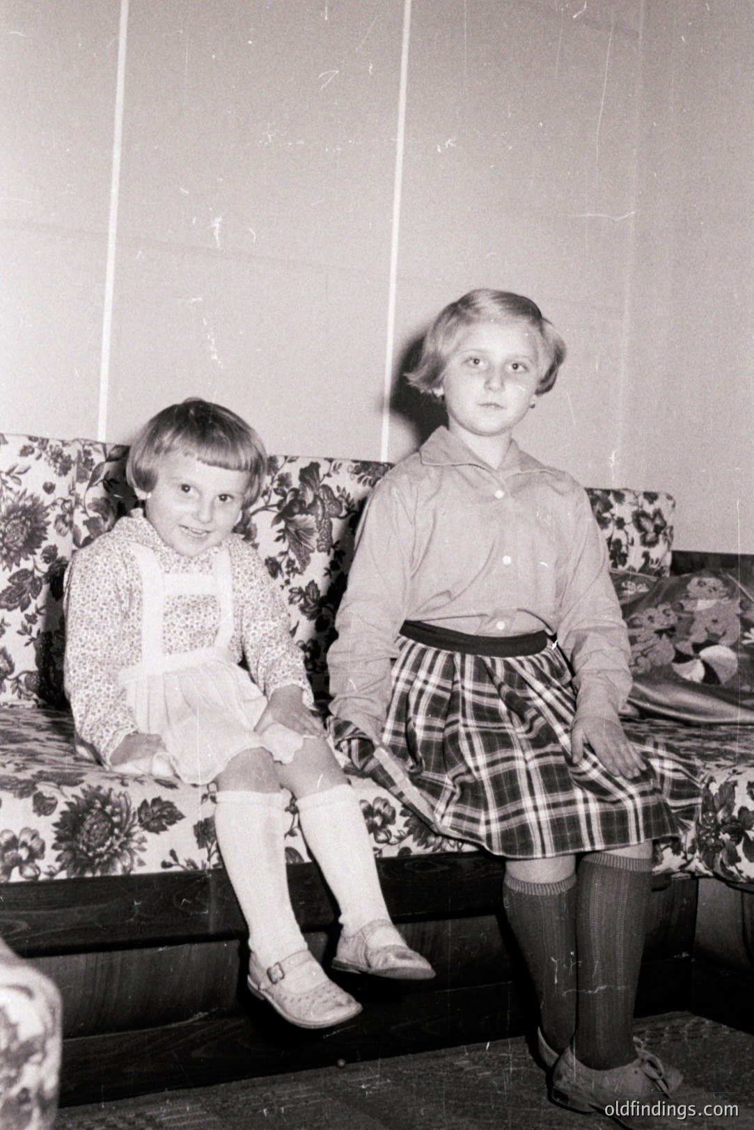 Two young girls, seated on a floral upholstered bench, pose for a formal portrait. Both wear dresses and knee-high socks; one has a Peter Pan collar. Interior setting; paneled walls visible. Likely a studio or formal home space, 1960s-1970s. Possible family archival photograph.