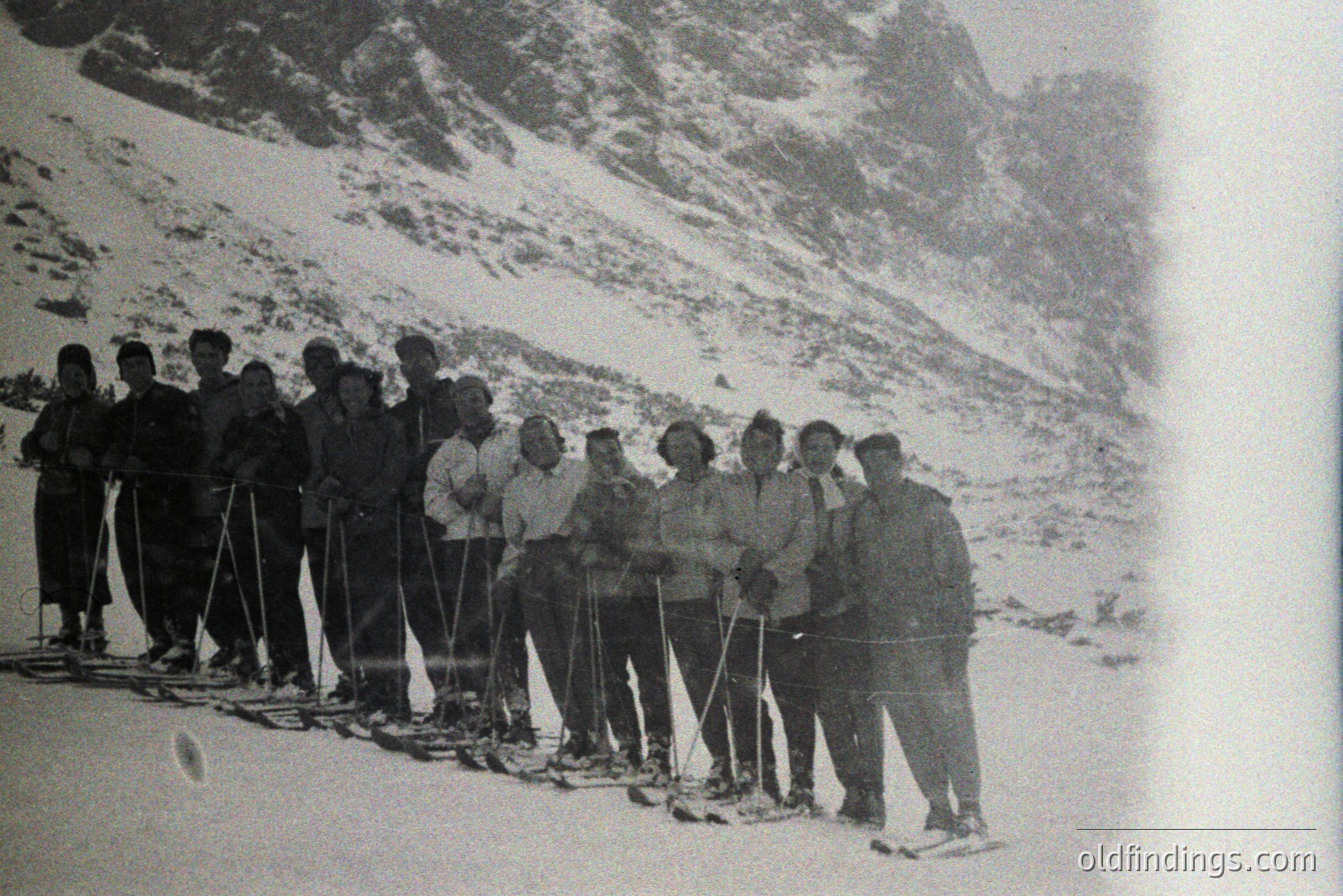 Group portrait of thirteen individuals, likely skiers, posed on a snow-covered slope with a dramatic mountain backdrop. Clothing suggests a mid-20th century timeframe (1950s-1970s). Traditional alpine attire is evident, possibly in a European location. Visible equipment includes skis and poles. A possible gathering of a ski club or expedition.