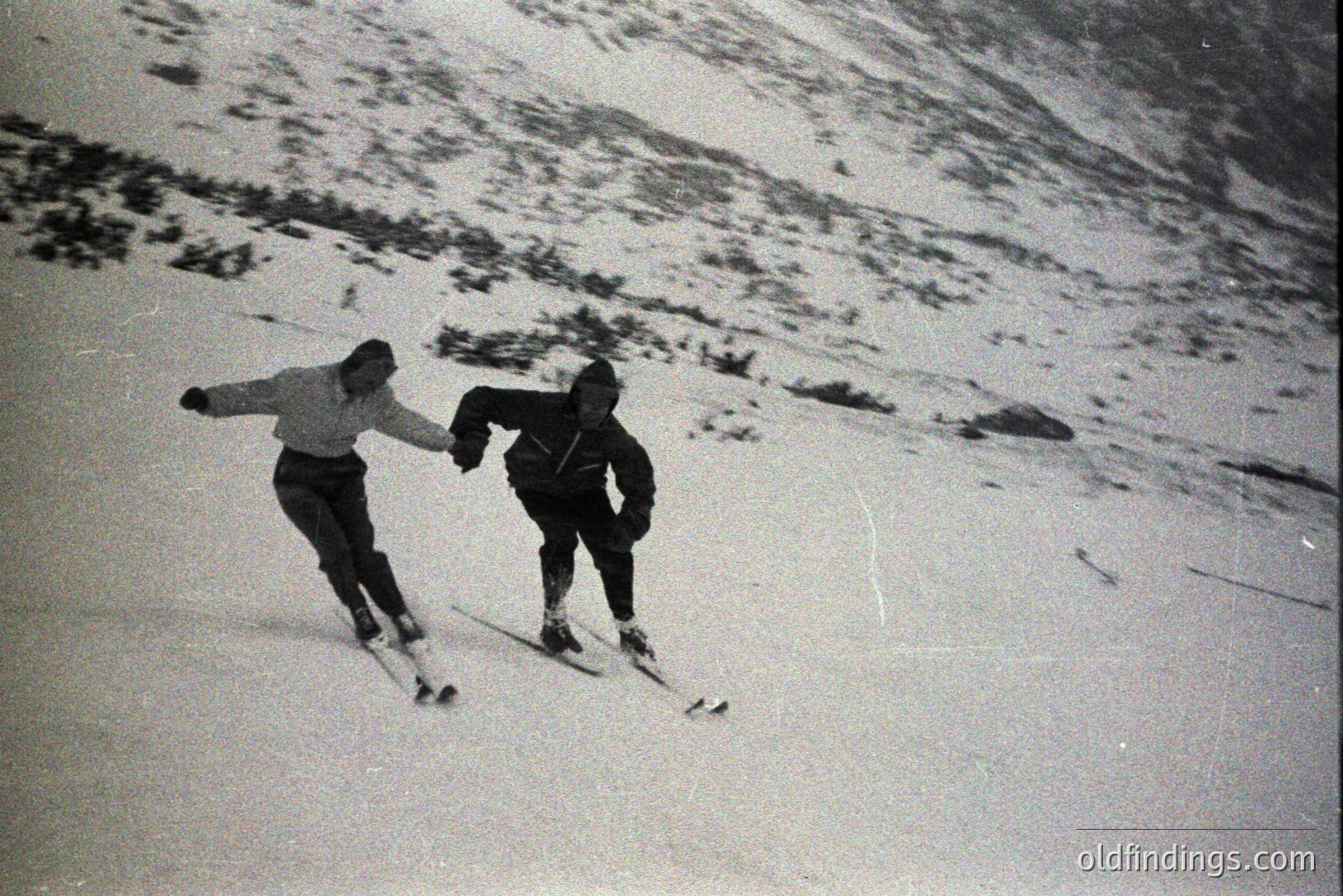 Two skiers descend a snow-covered slope, likely in an alpine region. The image, captured with a vintage film camera, exhibits grain and a shallow depth of field. Both skiers are dressed in what appear to be 1960s-era winter clothing. A hillside flanks the slope. Stock potential for vintage recreation imagery.