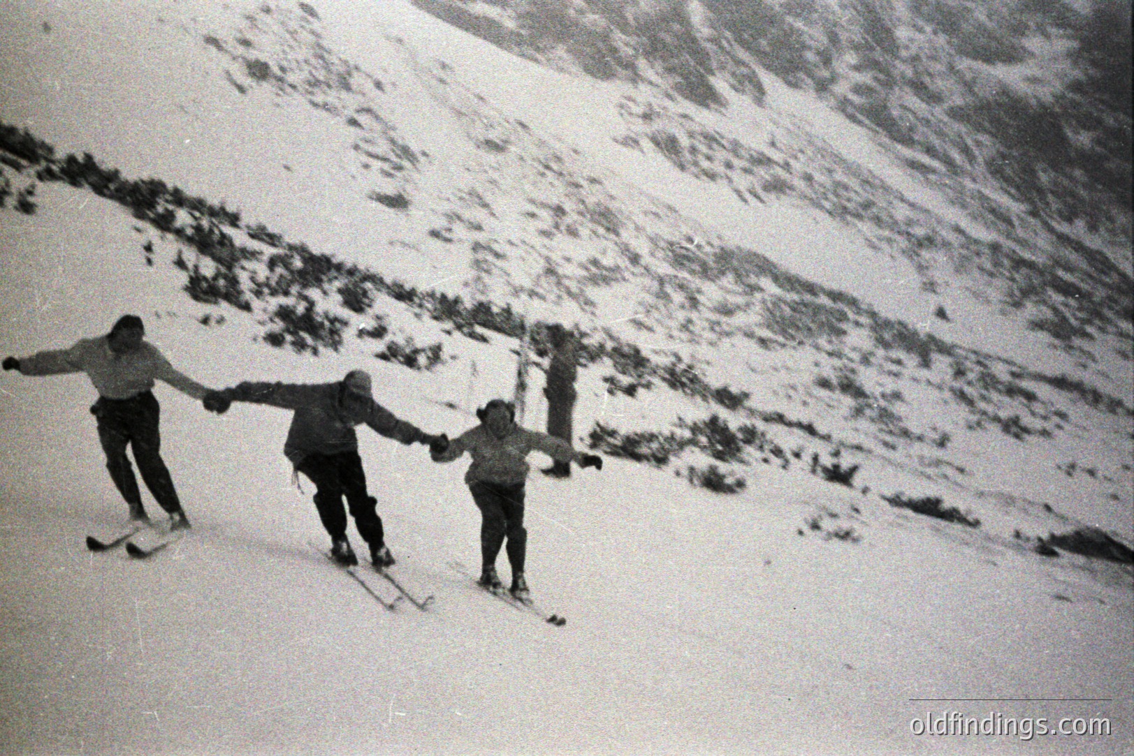 Three skiers descend a snow-covered slope. Visible clothing suggests a mid-century style. The backdrop features a snow-dusted mountain range, indicative of an alpine environment. Photo exhibits evident grain and tonal range characteristic of film. Likely a recreational activity snapshot.