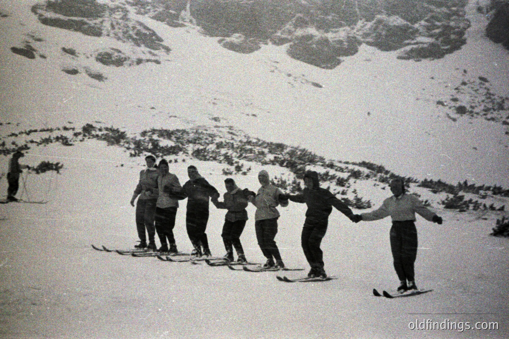 A group of seven people, all wearing winter clothing and standing on skis, hold hands in a circle on a snow-covered slope. Mountainous terrain fills the background, visible through falling snow. Likely a recreational winter activity scene, possibly 1950s or 60s.