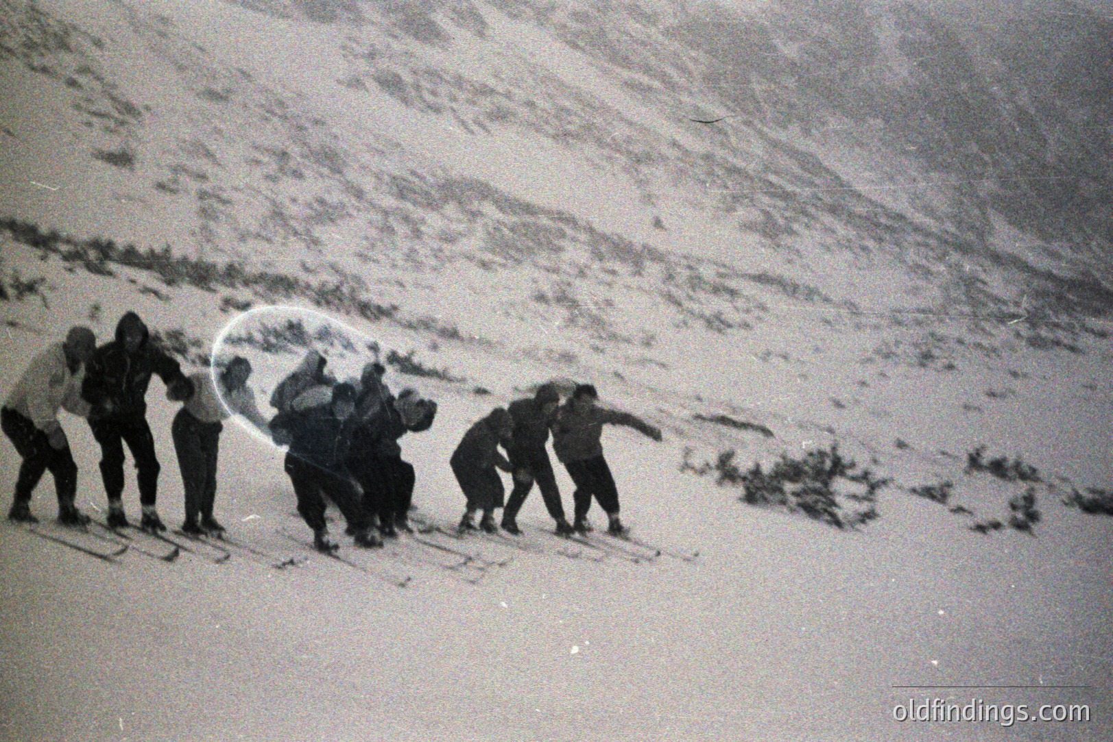 A group of skiers descends a steep, snow-covered slope. Figures appear bundled in dark clothing; the focus is somewhat blurred, suggesting movement. Likely a recreational or training scene. Appears to be from the mid-20th century. Possible location: mountainous region. Commercial value as stock footage or vintage design element.
