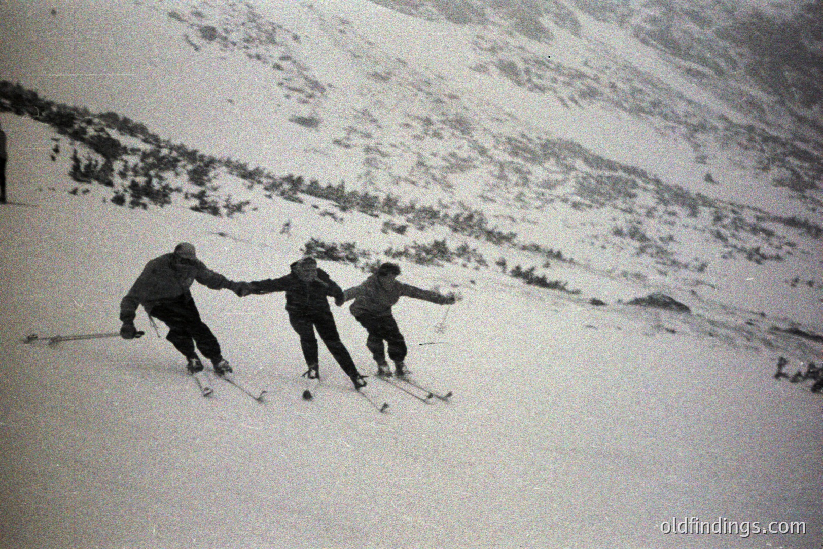 Three skiers descend a snow-covered slope. The individuals wear period clothing, with dark jackets and trousers. Visible terrain suggests an alpine environment. Likely a recreational winter scene, circa 1950s-1970s. A distinctive, slightly grainy photographic style.