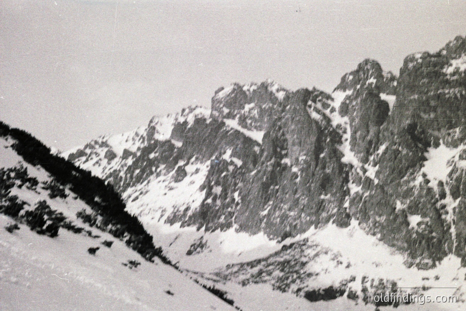 Dramatic, snow-covered alpine landscape featuring steep, rocky peaks. Sparse vegetation clings to the lower slopes. Likely a tourist view from the 1960s or 70s, exhibiting a classic documentary aesthetic. Potential for use in travel or design projects.