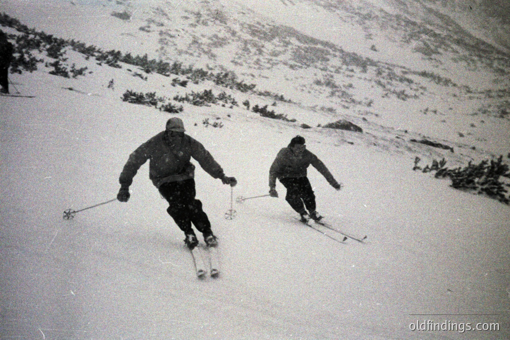 Two skiers descend a snow-covered slope, captured in a vintage black and white photograph. The men wear heavy winter clothing and hats, showcasing mid-century ski attire. Visible terrain suggests an alpine environment, possibly a mountainous resort. Likely 1950s-1970s. A stock photo reference for vintage winter scenes.