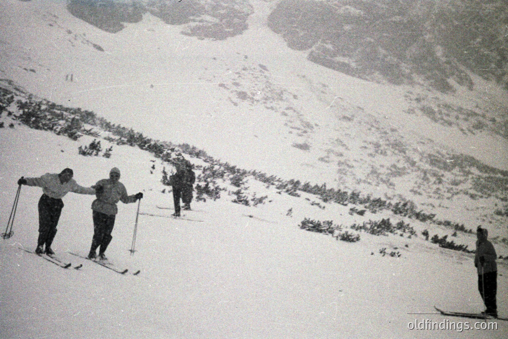 Four figures ski down a snow-covered slope, surrounded by pine forest and towering peaks. Likely taken in the mid-20th century, the skiers are outfitted in vintage snow gear. The image evokes a sense of adventure in a mountainous landscape.