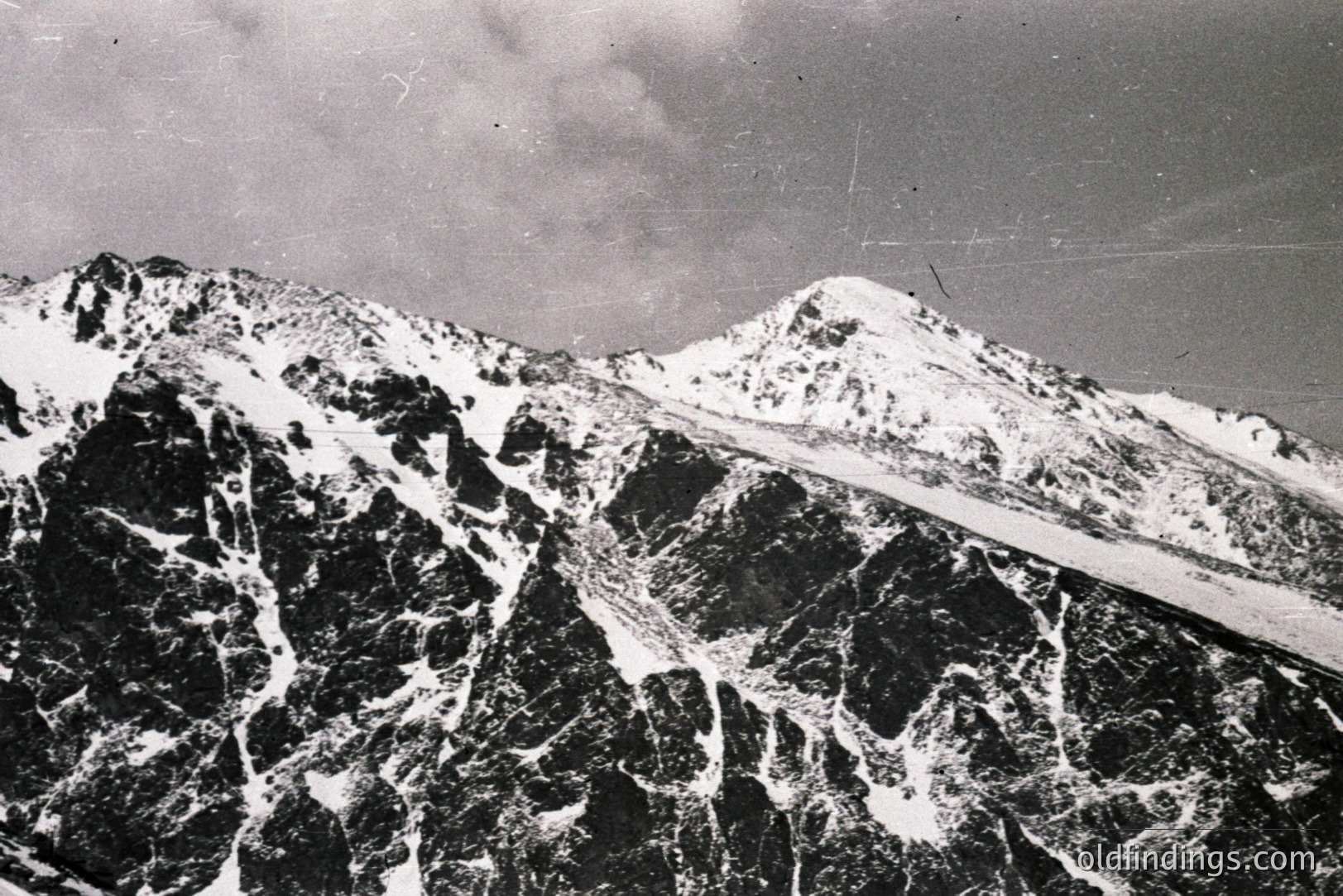 Dramatic, snow-covered peaks dominate the landscape. Stark contrast highlights a rugged alpine terrain with visible rock formations and glacial ice. The image has a grainy quality, indicative of an older photographic process. Likely a mid-20th century scenic view.