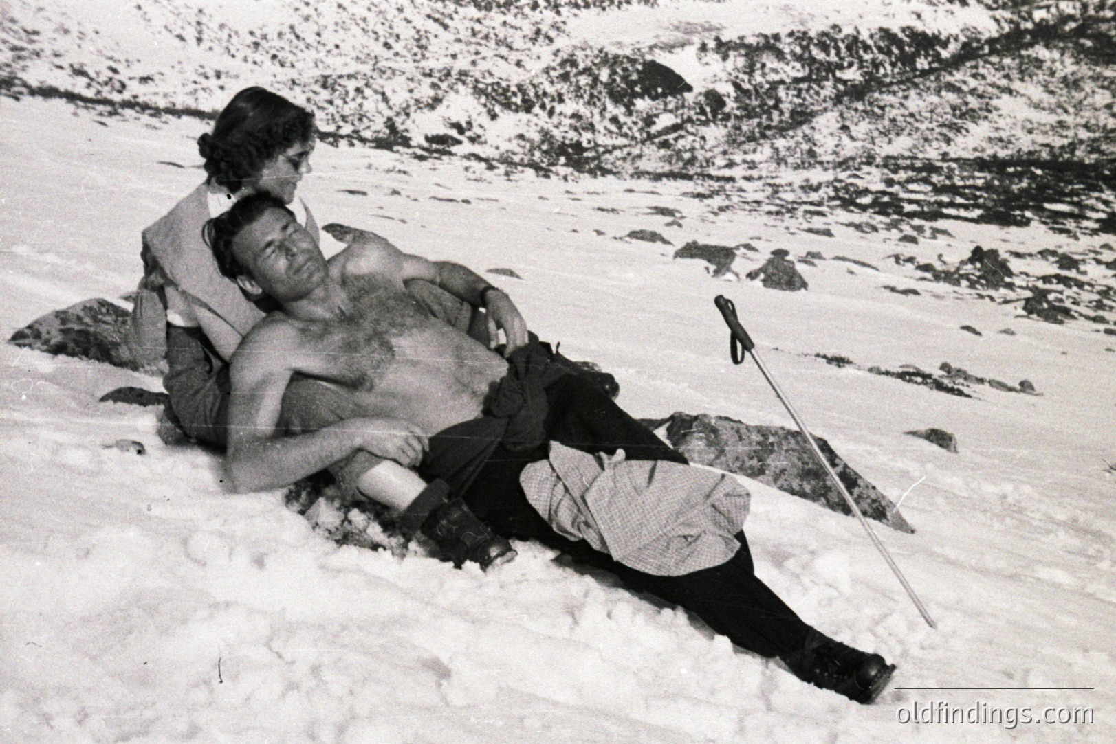 Man lies prone in snow, partially unclothed, supported by a woman. Likely an alpine environment, judging by the snow-covered peaks in the background. Possible mountaineering accident or staged scene. Vintage photographic style, likely 1930s-1950s. Image may hold commercial value for historical stock or design.