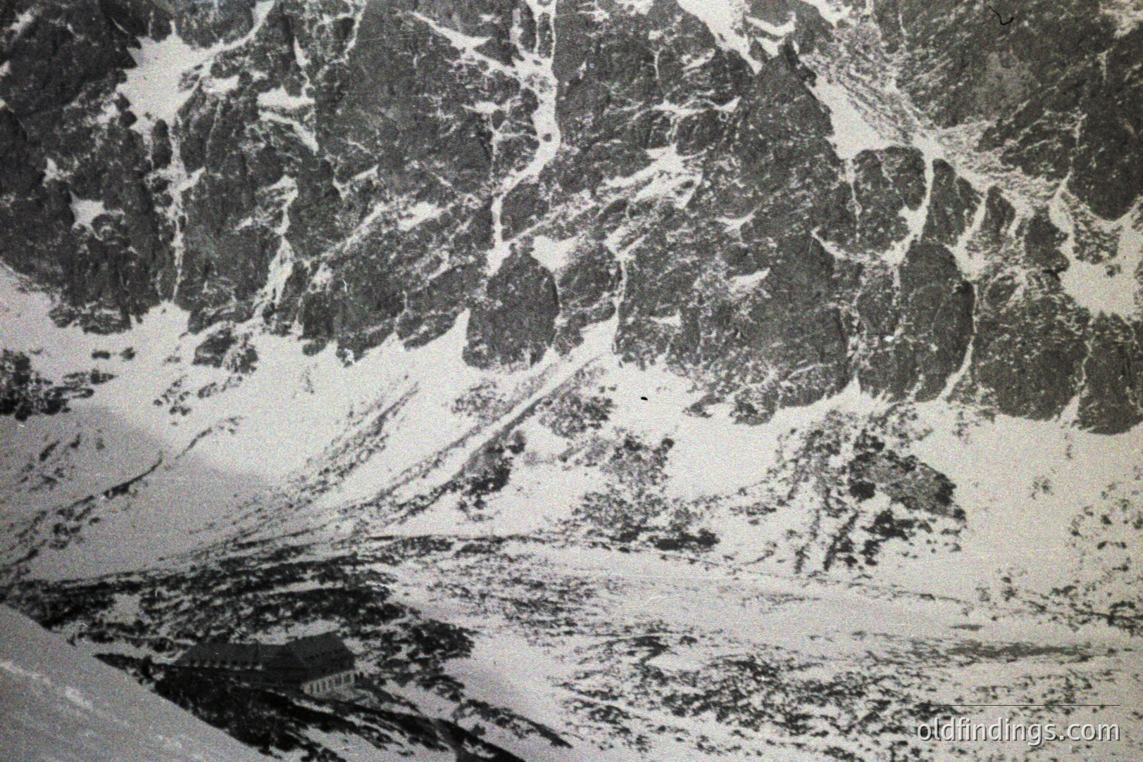 A high-angle, monochrome photograph reveals a snow-covered alpine landscape. Jagged peaks dominate, interspersed with deep valleys and drifts. A small structure is visible at the base of the slope. Likely taken in the mid-20th century, showcasing glacial terrain. Potential value for research on alpine geology or historical mountaineering.