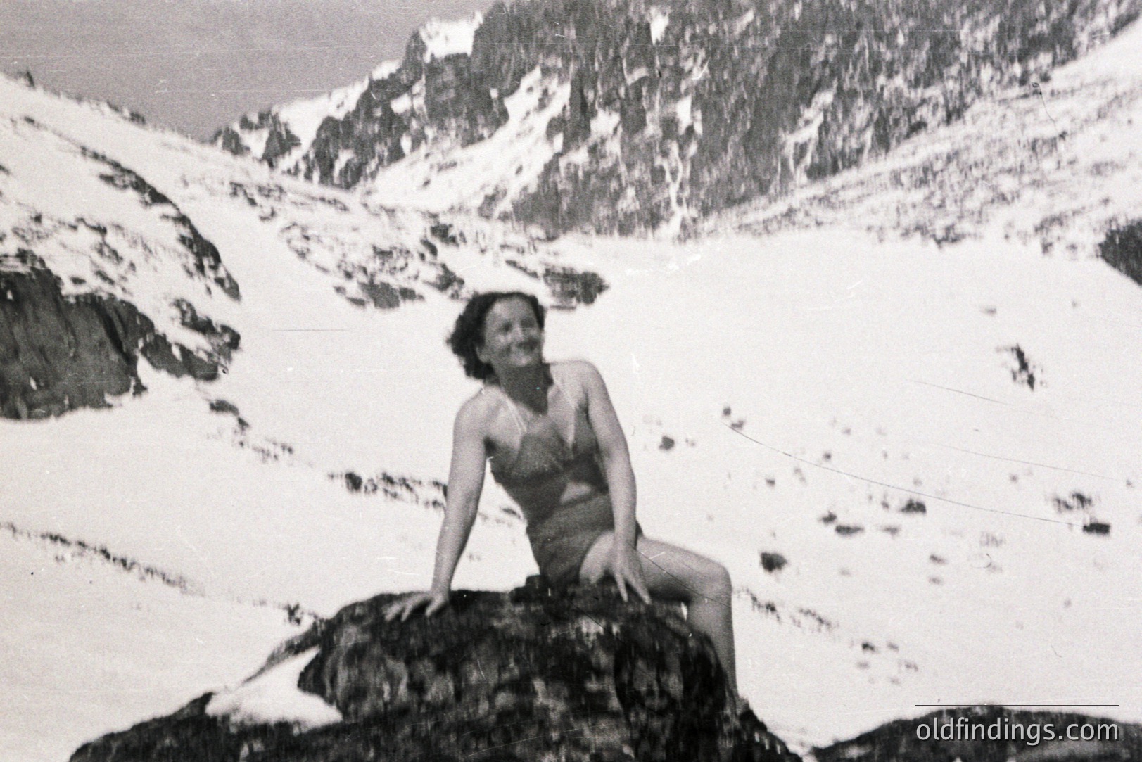 A young woman in a vintage swimsuit sits perched on a rock amidst a snowy, mountainous landscape. Likely a promotional shot showcasing swimwear or outdoor leisure. The stark contrast and composition suggest a 1960s or 70s origin. The backdrop features rugged, snow-covered peaks.