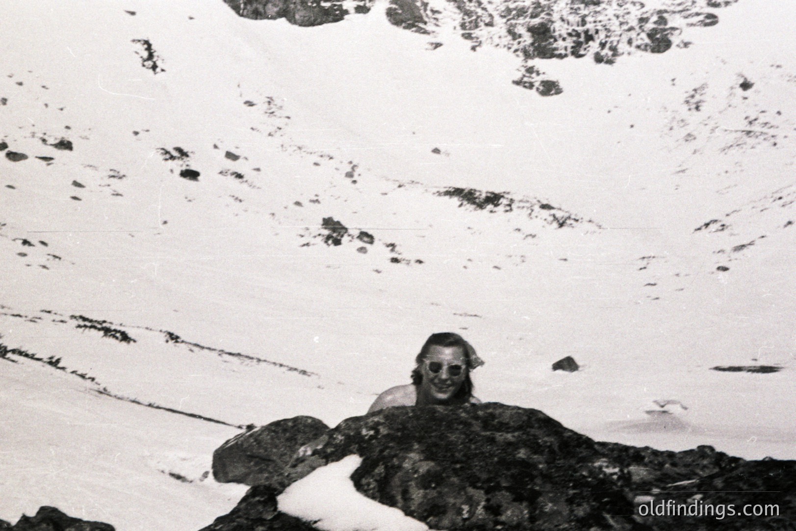 A man with long hair smiles directly at the camera while partially sheltered by dark rocks covered in snow. Sparse vegetation appears in the background. Likely a candid outdoor portrait, possibly an alpine region. Estimated 1970s aesthetic. Appears suitable for historical context or design.