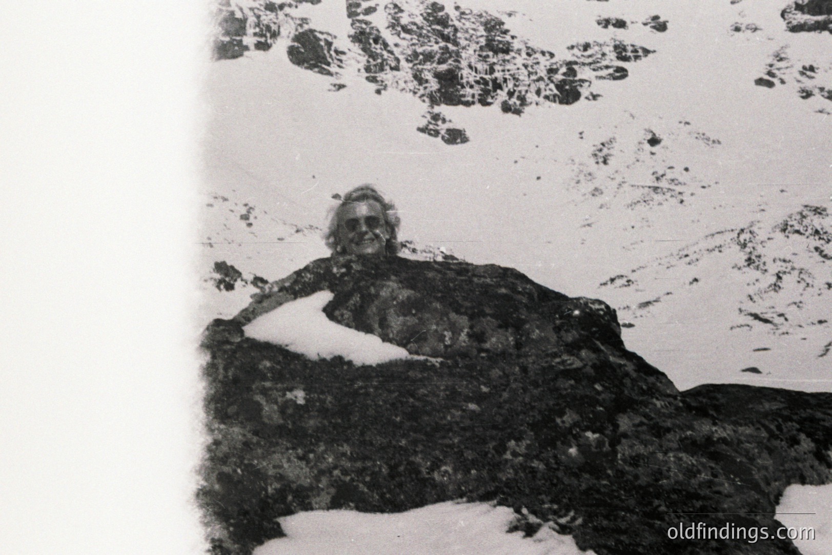 A man with a full beard sits atop a snow-covered boulder, gazing toward a mountainous, snowy landscape. The image, likely a personal snapshot, evokes a rugged, outdoor lifestyle. Grainy texture suggests older film stock, possibly 1970s or 80s. Location undetermined. Photo's aesthetic could be useful for vintage design.