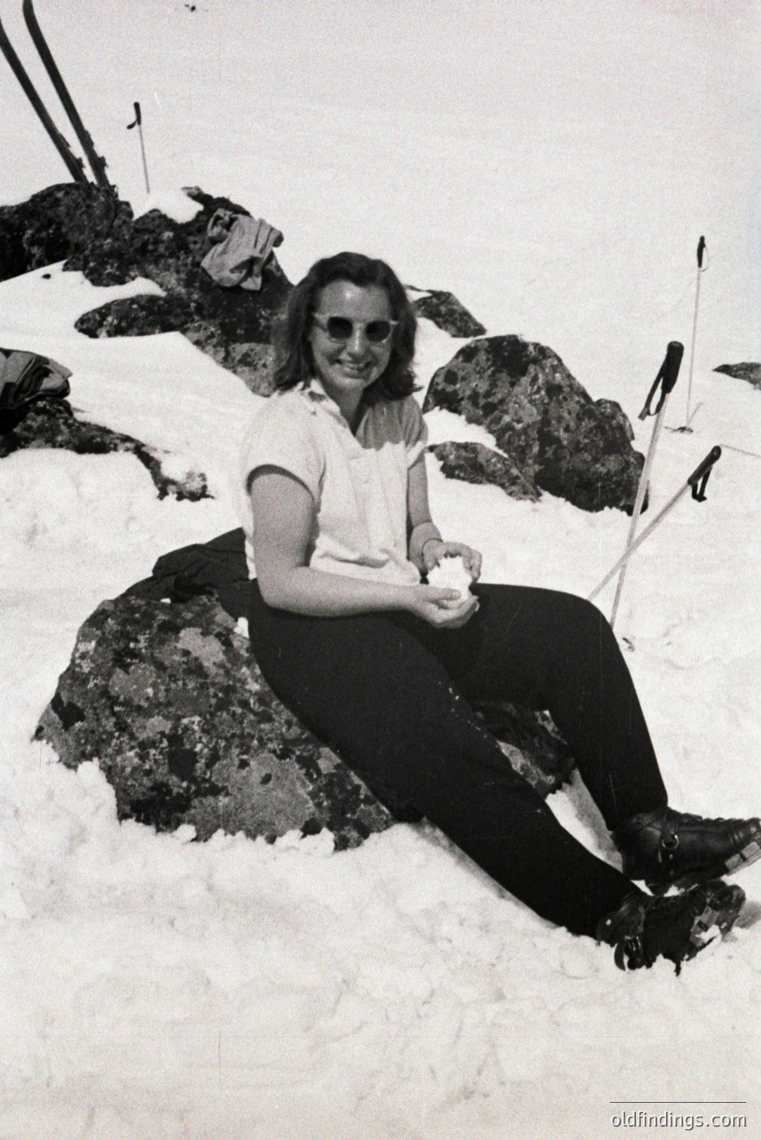 A woman sits on a boulder amidst a snowy landscape, wearing a short-sleeved white shirt, dark trousers, and sunglasses. Likely a winter recreation scene, possibly skiing or hiking. The style and clothing suggest a 1960s-1970s timeframe. A snow ball is held in her gloved hand.