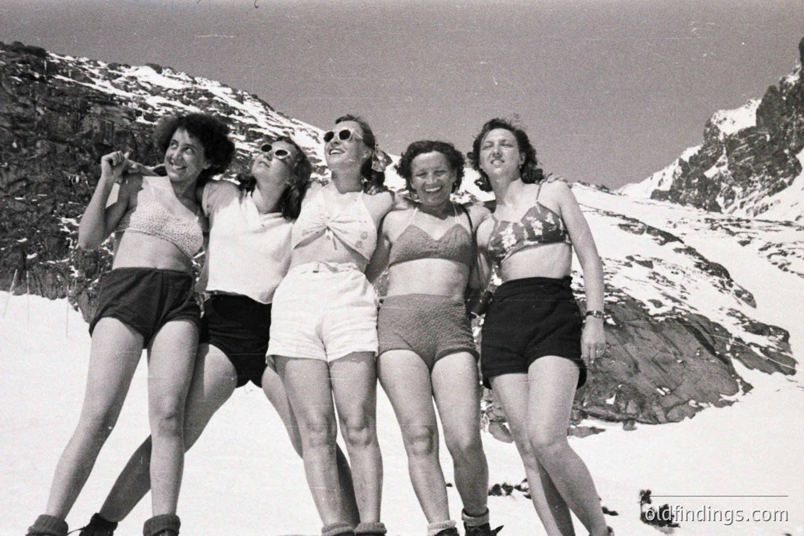 Five women stand outdoors, bundled in swimwear and posing against a snowy mountain backdrop. Vintage style knickers and bandeaus suggest a 1950s or 1960s leisure scene. Likely captured during a winter sports holiday. Commercial value for fashion or historical research.