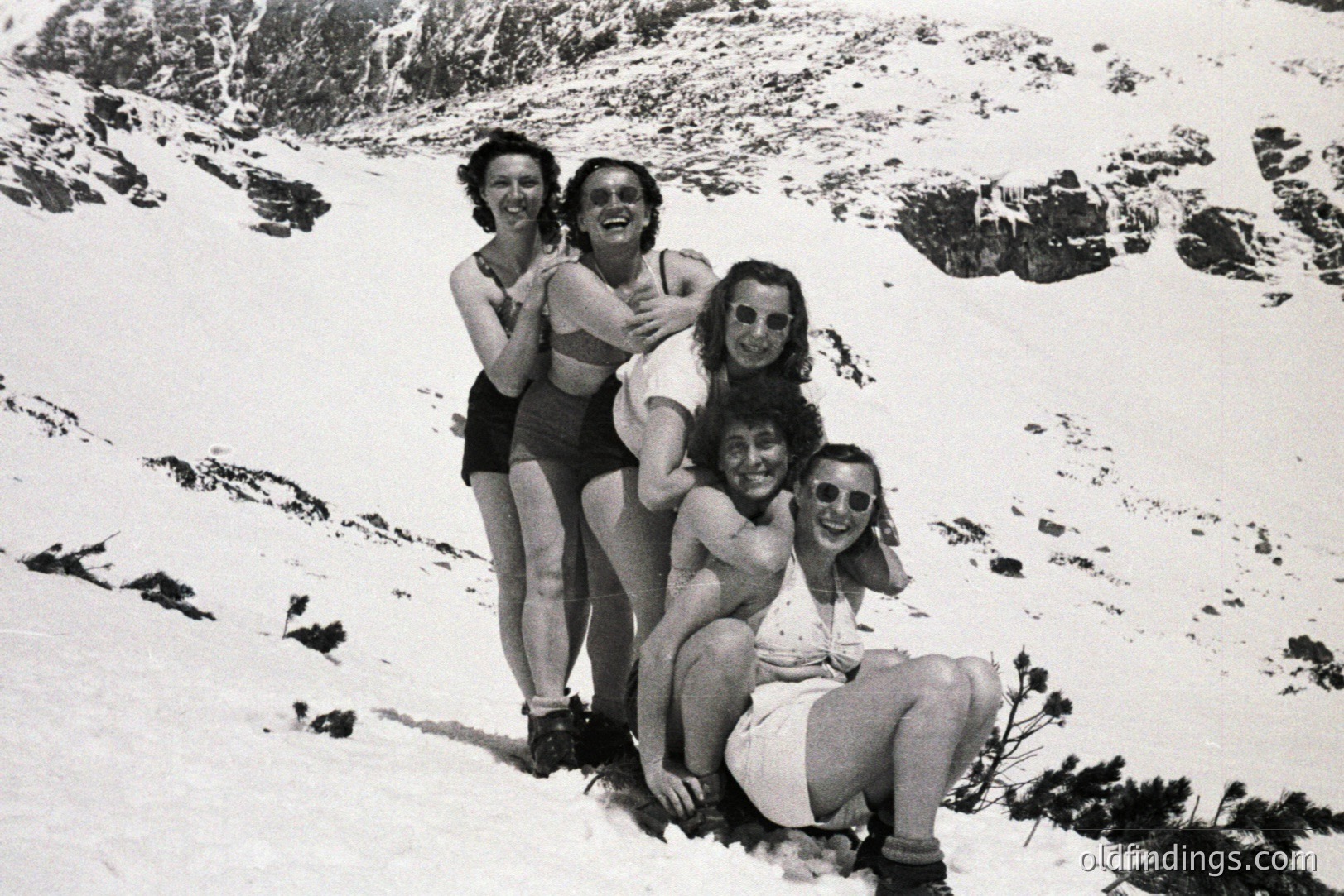A group of five people in swimwear poses in a snowy, alpine landscape. All wear sunglasses and appear joyful. The composition, lighting, and clothing suggest a mid-20th century travel or leisure photograph, possibly promotional. Location is unconfirmed.