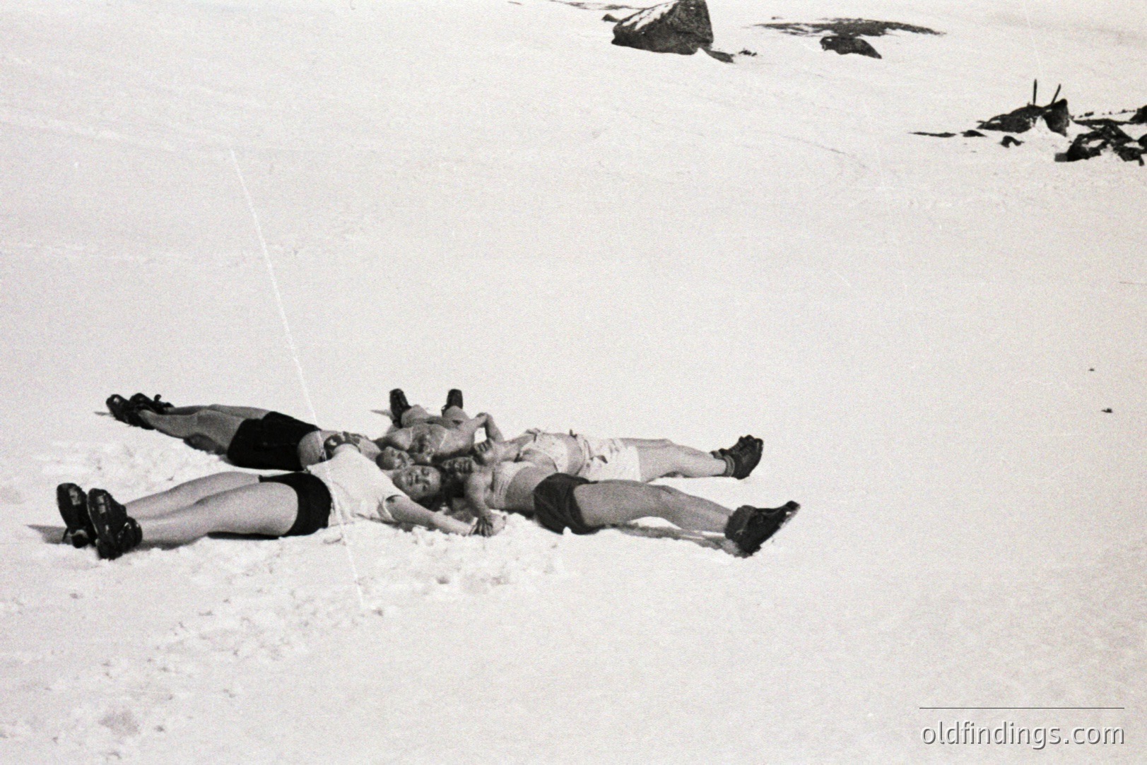 Three figures lie intertwined in a snowdrift, appearing relaxed or playfully entangled. Their attire - shorts & boots - suggests a casual, outdoor recreation. The monochrome aesthetic and photographic style indicate a likely mid-20th century origin. Possible alpine or mountain environment.