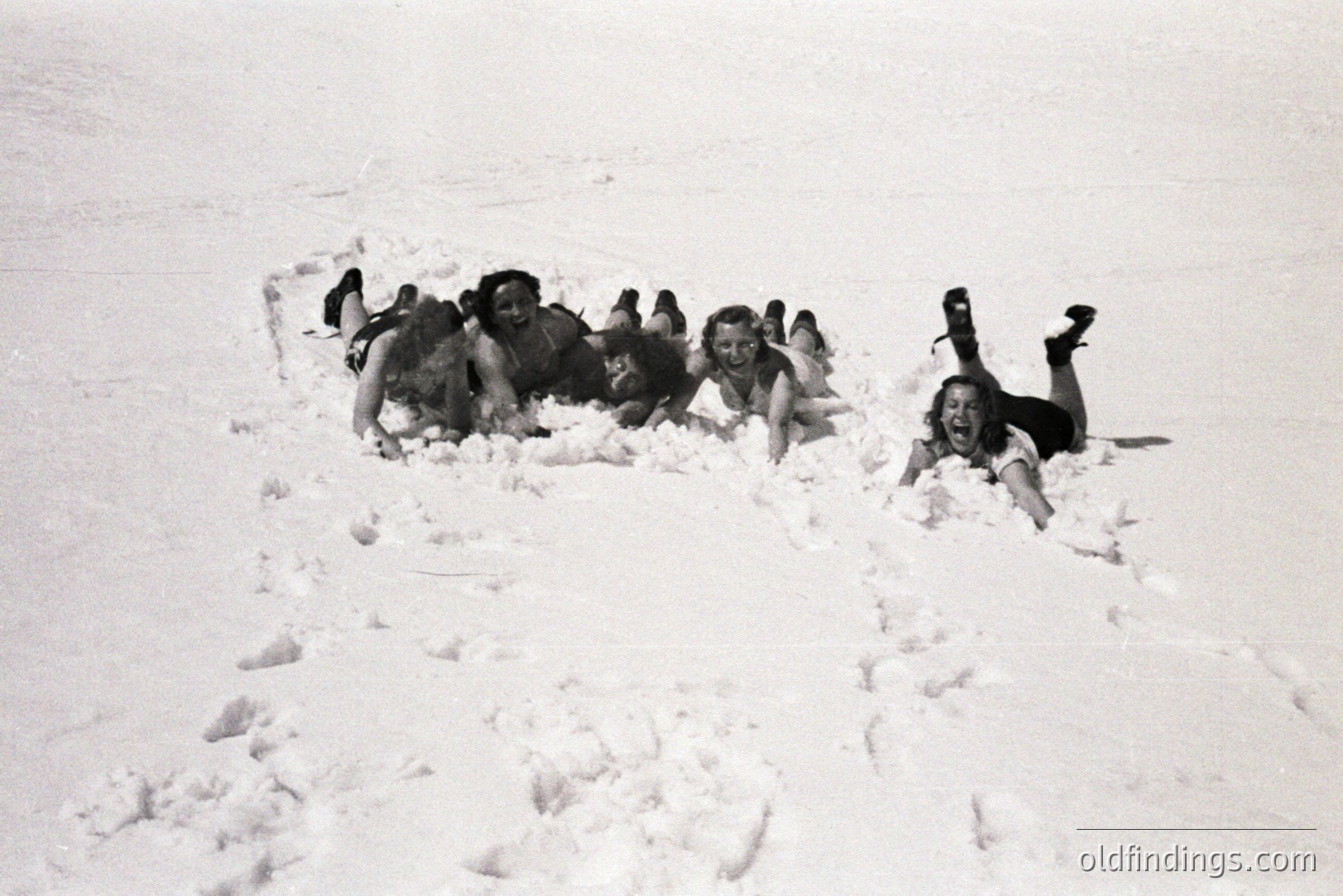 Four figures lie playfully in a snow-covered landscape, appearing to be enjoying a moment of winter recreation. Clothing suggests a casual style of the mid-20th century. The scene conveys a sense of youthful exuberance and snowy leisure. Appears to be a candid snapshot.