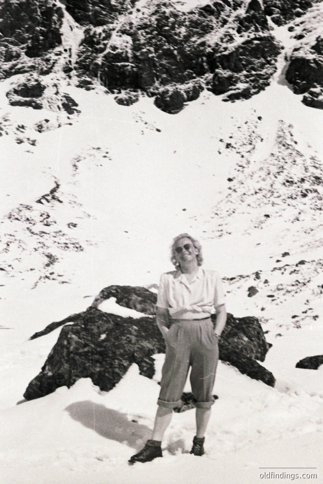A woman in 1960s-era clothing stands confidently amidst a snowy, rocky alpine landscape. She wears wide-leg trousers, a button-down shirt, and dark boots. Visible rock formations and expansive snow suggest a high-altitude location. An outdoor portrait, possibly for travel or adventure documentation.